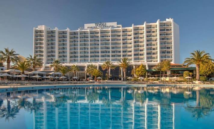 Large hotel with the sign 'Tivoli Marina Vilamoura' on the top, overlooking a swimming pool with clear blue water, surrounded by palm trees and outdoor seating area.