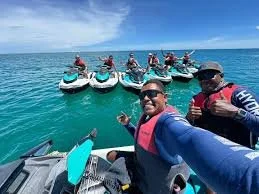 Group of people taking a selfie near a boat with jet skis on a clear, blue ocean with a sunny sky.
