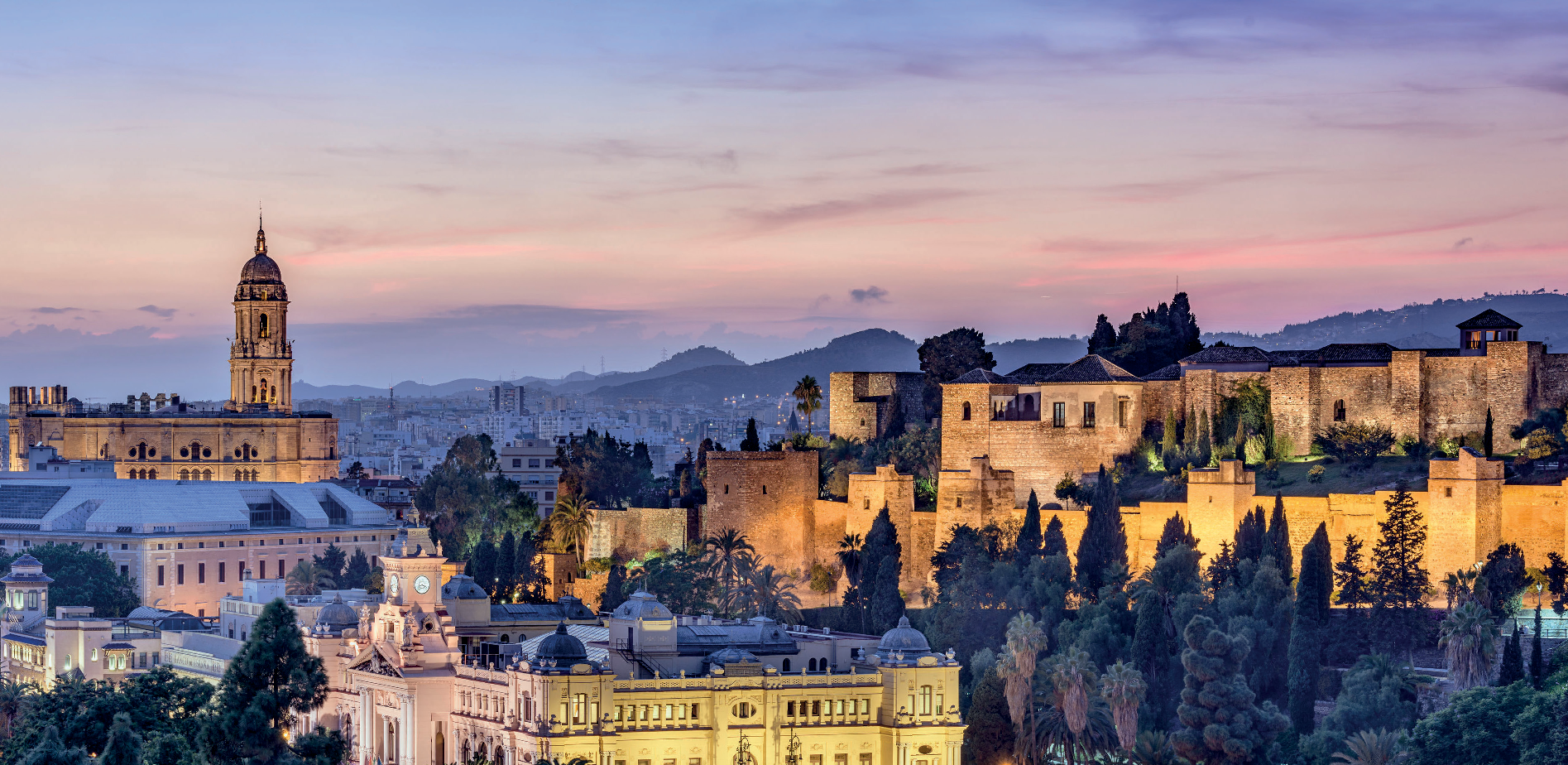 Cityscape at dusk with historical buildings, a castle wall, and a church tower against a backdrop of mountains and a colorful sky.