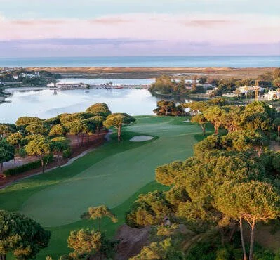 A golf course with neatly trimmed greens and trees, overlooking a body of water and a distant shoreline under a partly cloudy sky.