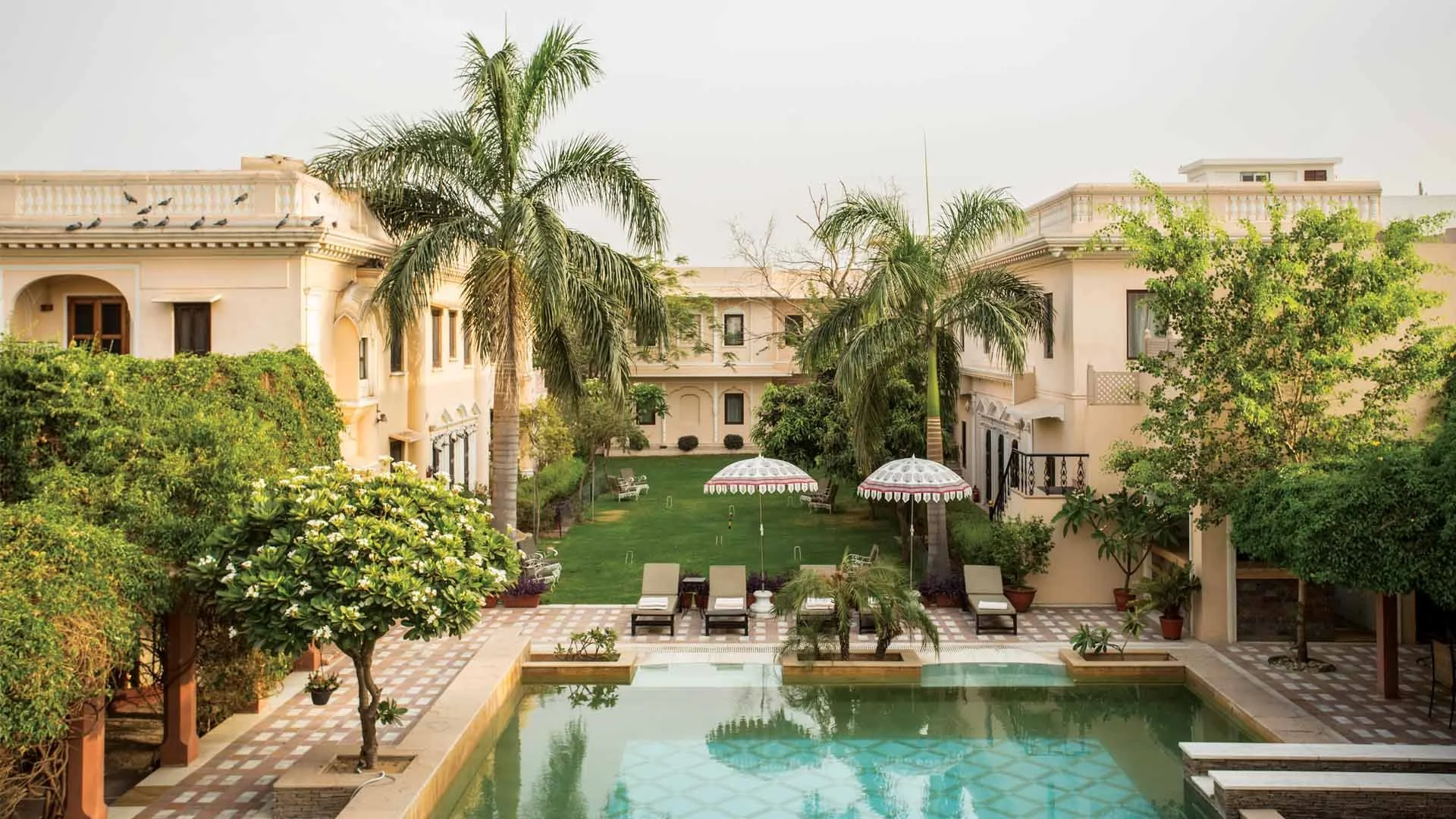 View of a luxurious residential property with a central courtyard featuring a swimming pool, lounge chairs, umbrellas, lush green trees, palm trees, and a grand building with balconies and ornate architectural details.