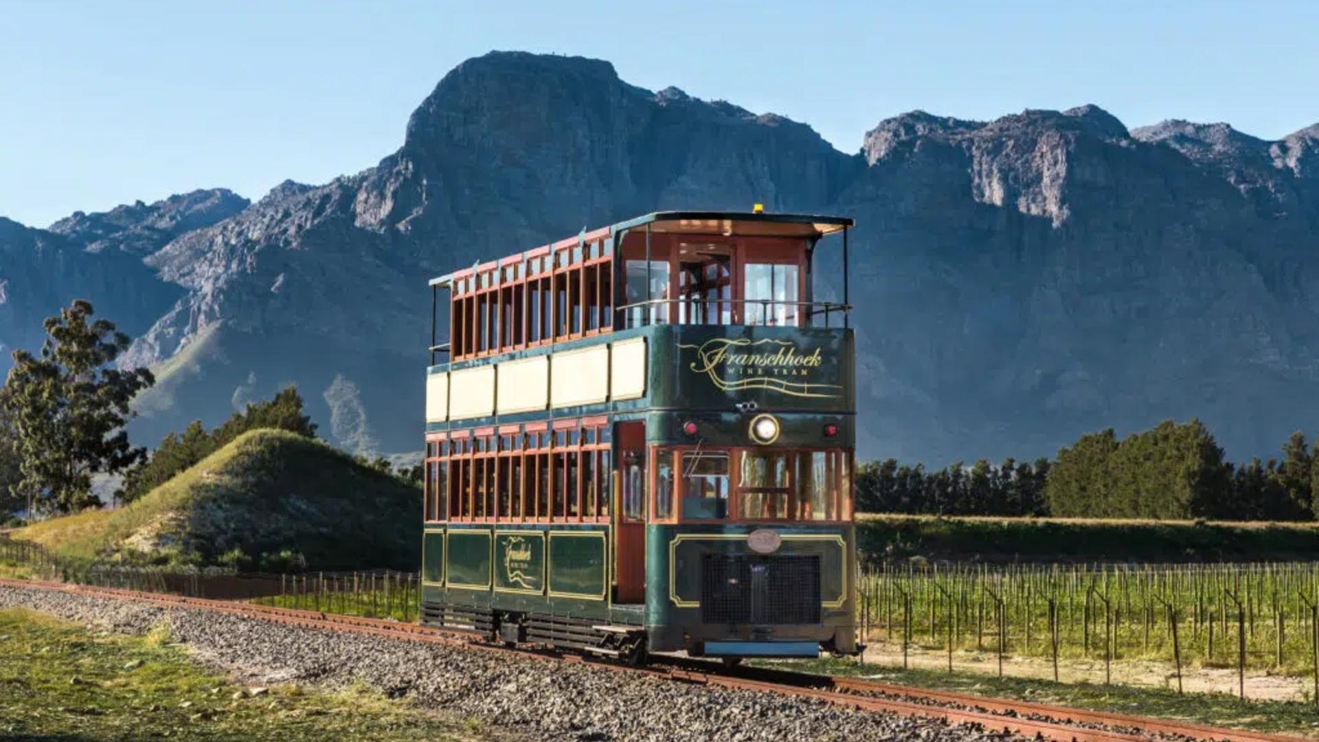Vintage double-decker tram on railway tracks with Green and red colors, set against a backdrop of tall mountain peaks and lush greenery.