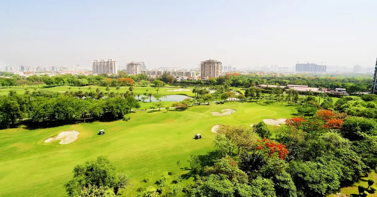 A scenic view of a golf course with sand traps, green fairways, and a water hazard, surrounded by trees and buildings in the background.