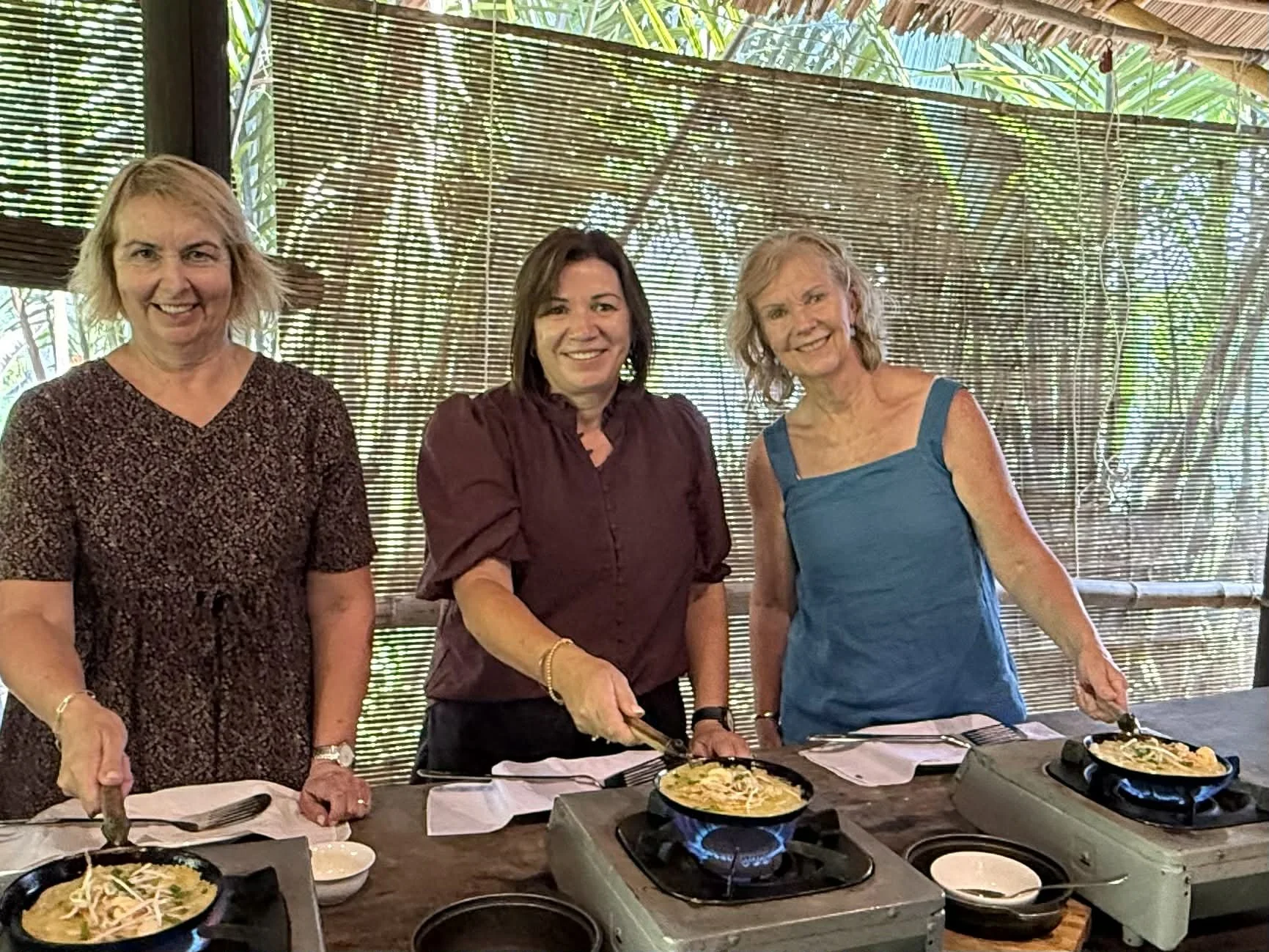 Three women cooking at a restaurant or kitchen, two are frying food on portable stoves, surrounded by cooking utensils and ingredients. They are smiling and appear to be enjoying the activity.