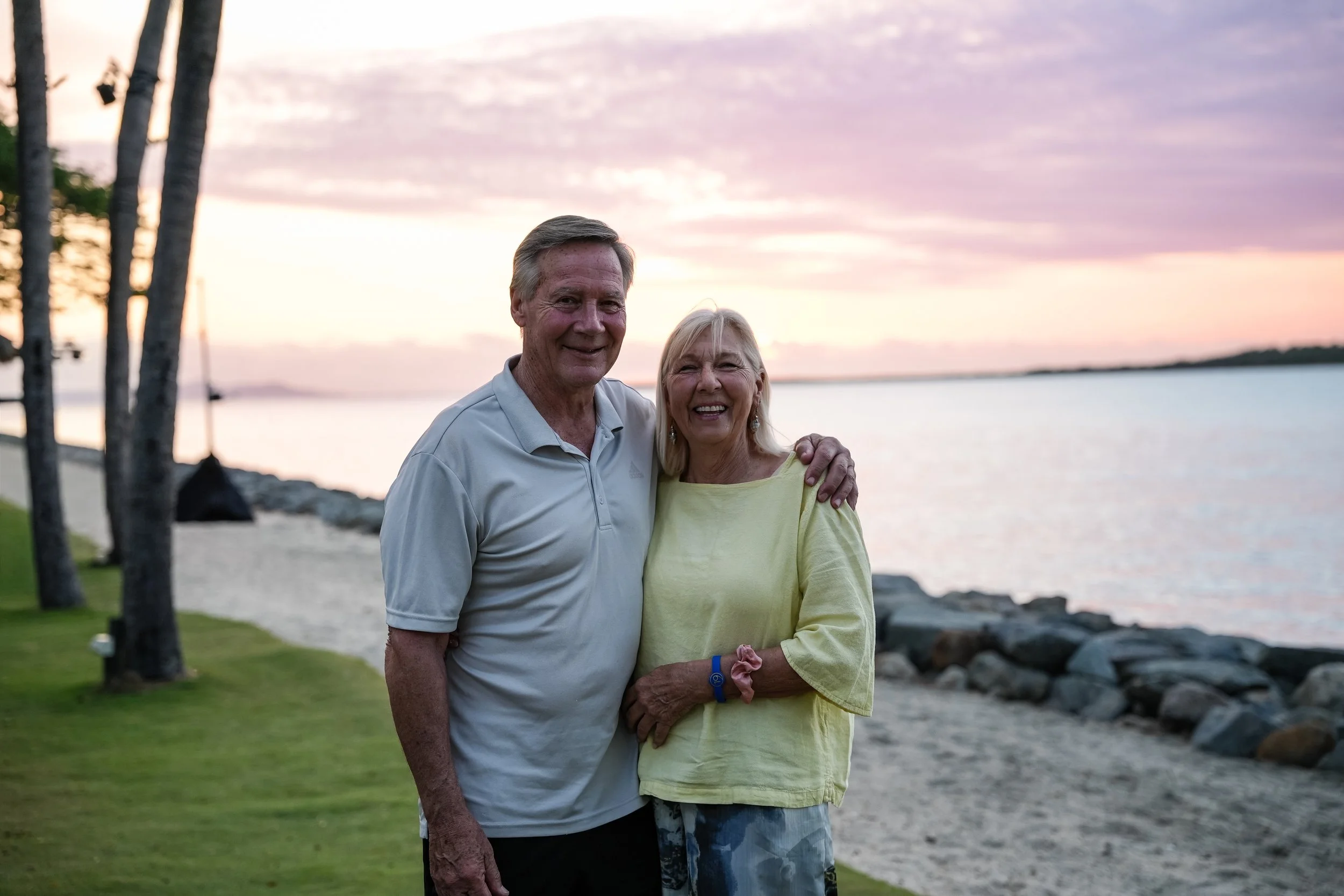 An elderly couple standing close together on a lakeside promenade during sunset, smiling at the camera with trees and water in the background.