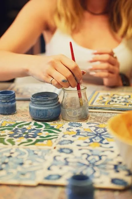 Woman painting on a ceramic tile with blue paint using a small brush, surrounded by jars of blue paint and a colorful tile pattern on the table.
