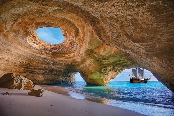 View of a cave at the beach with a large opening in the ceiling, showcasing the sky, and a ship sailing near the shoreline.