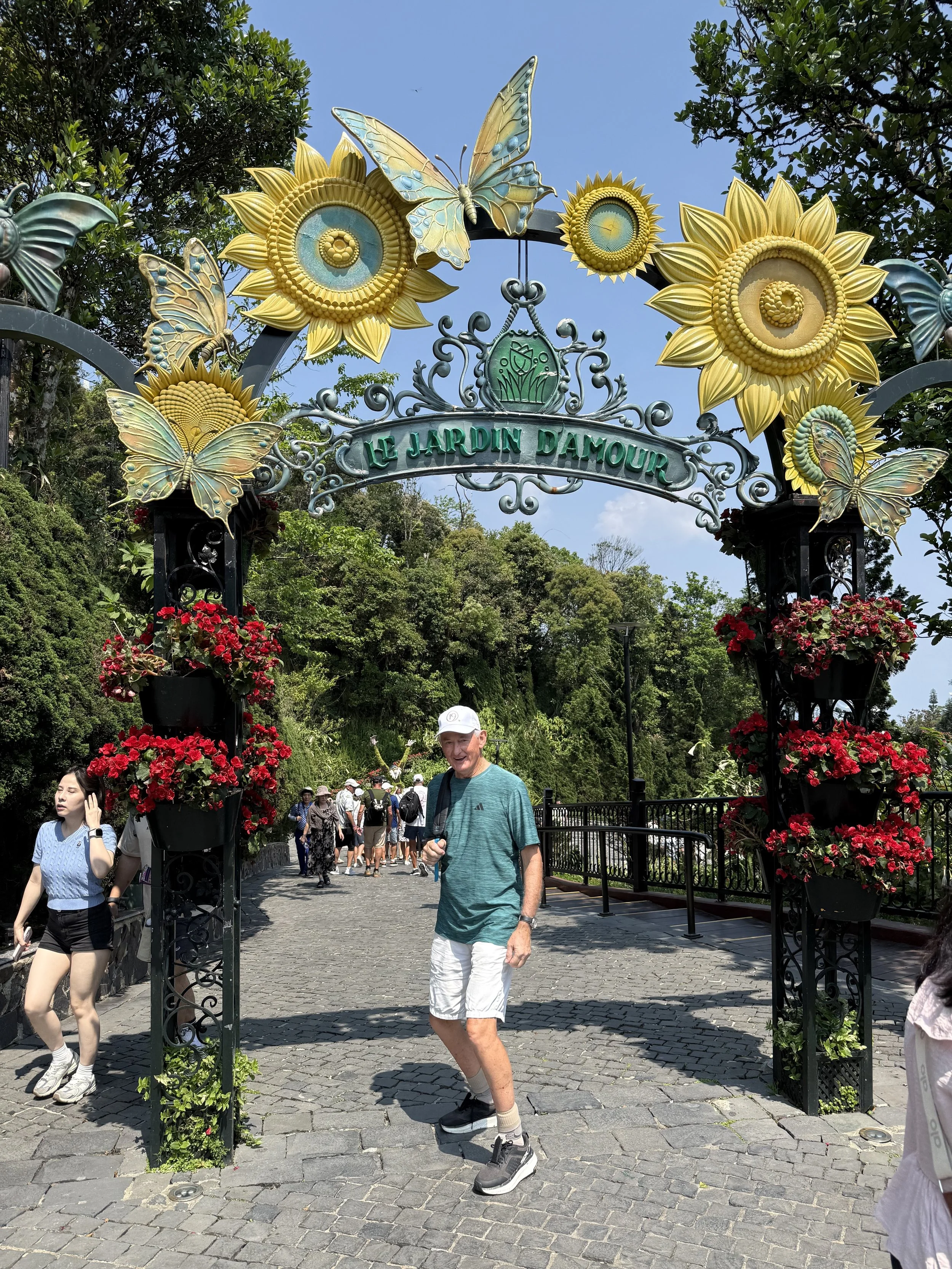 Entrance archway at Le Jardin D’amour with large sunflowers and butterflies, with people walking through on a paved path, surrounded by greenery and flowers.