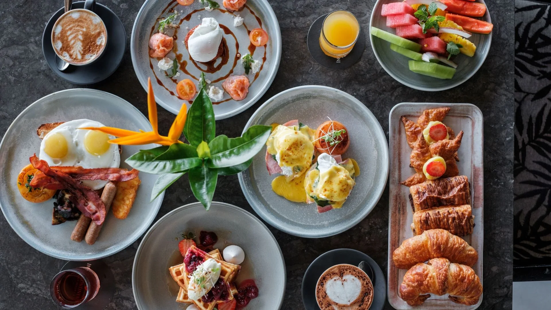 A variety of breakfast dishes on a dark marble table, including eggs Benedict, waffles, croissants, fresh fruit, and coffee.