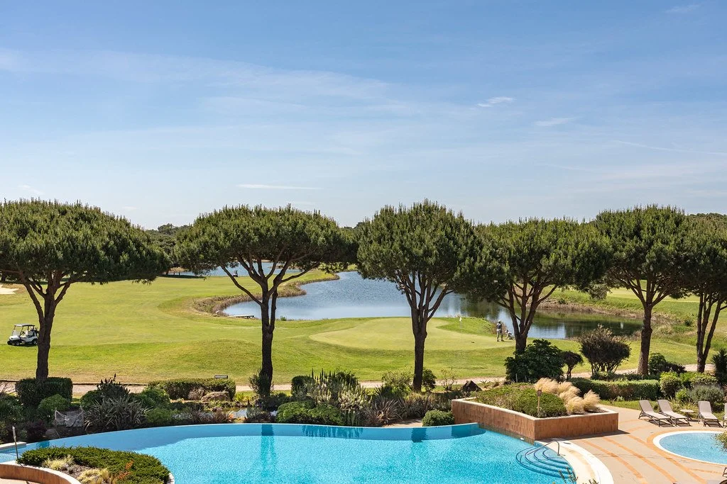 Golf course with trees, a pond, and a player near the water, viewed from a poolside area.