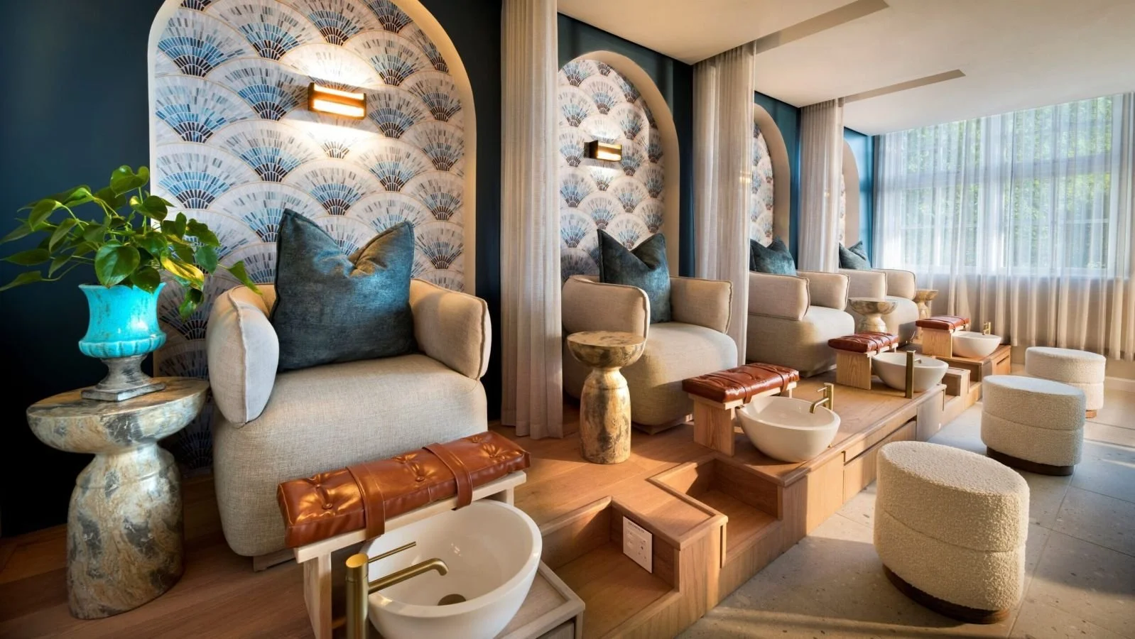 A modern salon with four massage chairs, each with a large dark blue cushion, illuminated by wall sconces, and a large window with sheer curtains allowing natural light. There are small wooden tables with foot baths, potted plants, and decorative wallpaper with fan patterns.
