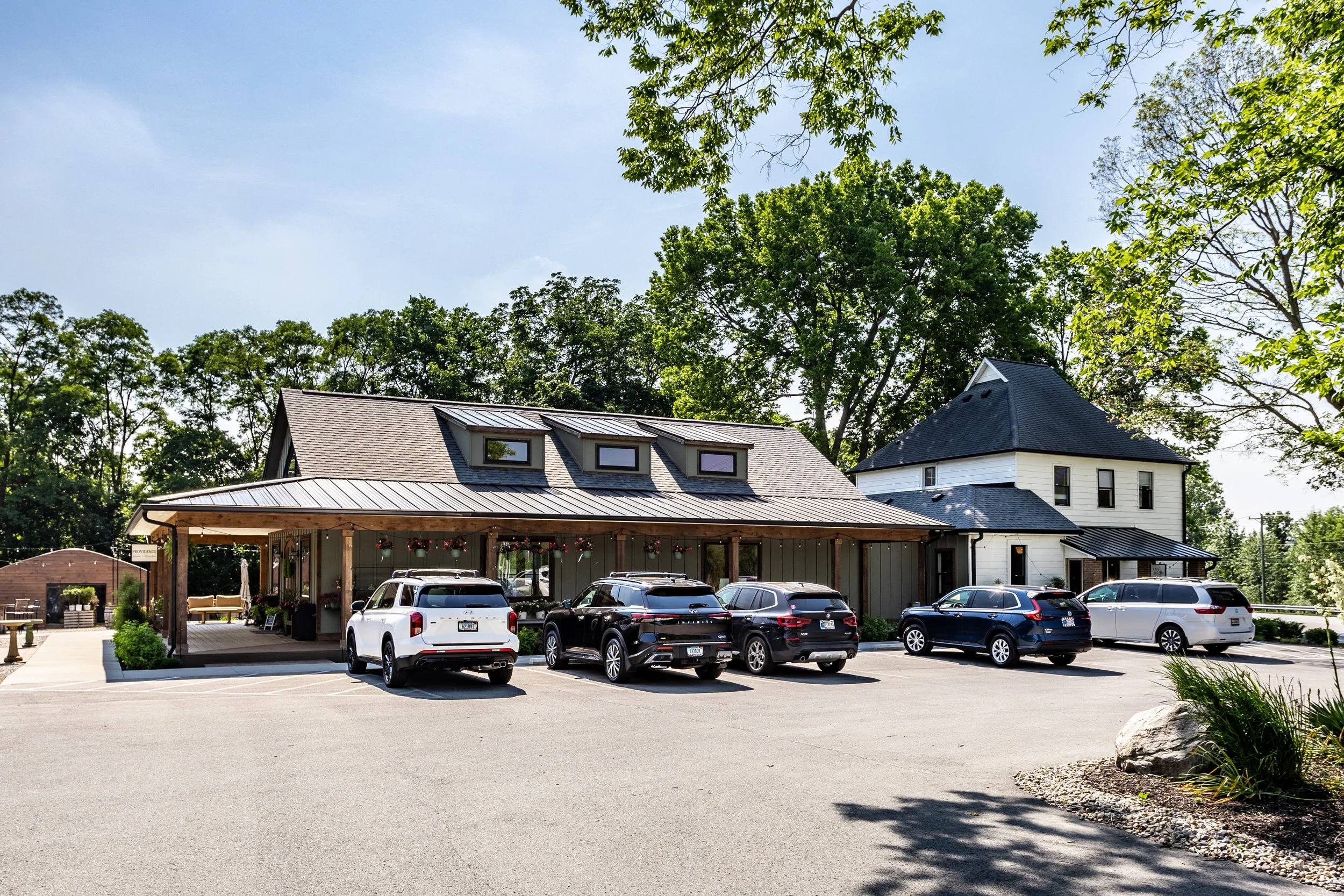 A parking lot in front of a modern building with a covered patio area, five parked cars, surrounded by green trees and landscaping.