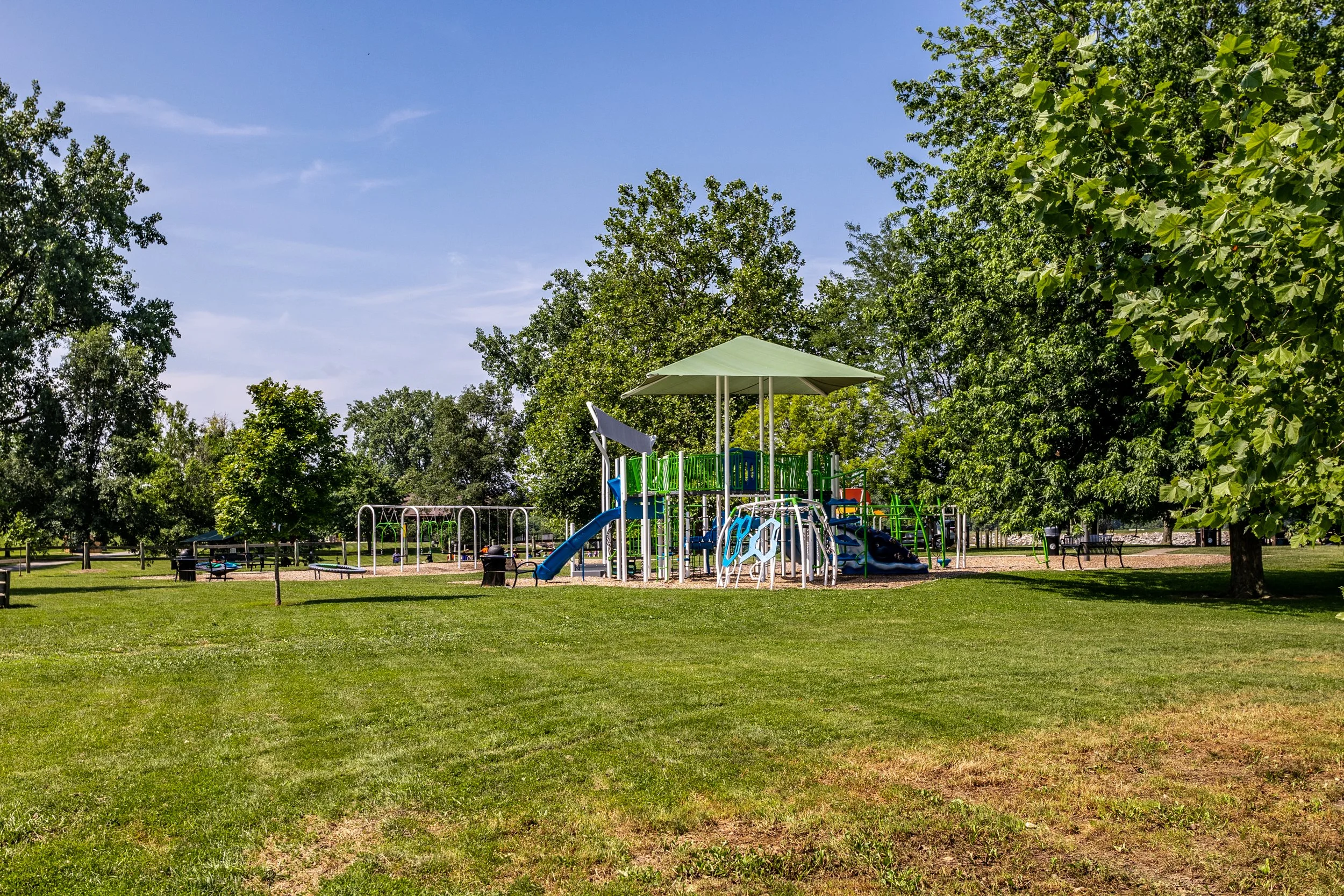 Children's playground with slides, climbing structures, and swings in a park under a clear blue sky, surrounded by green trees.
