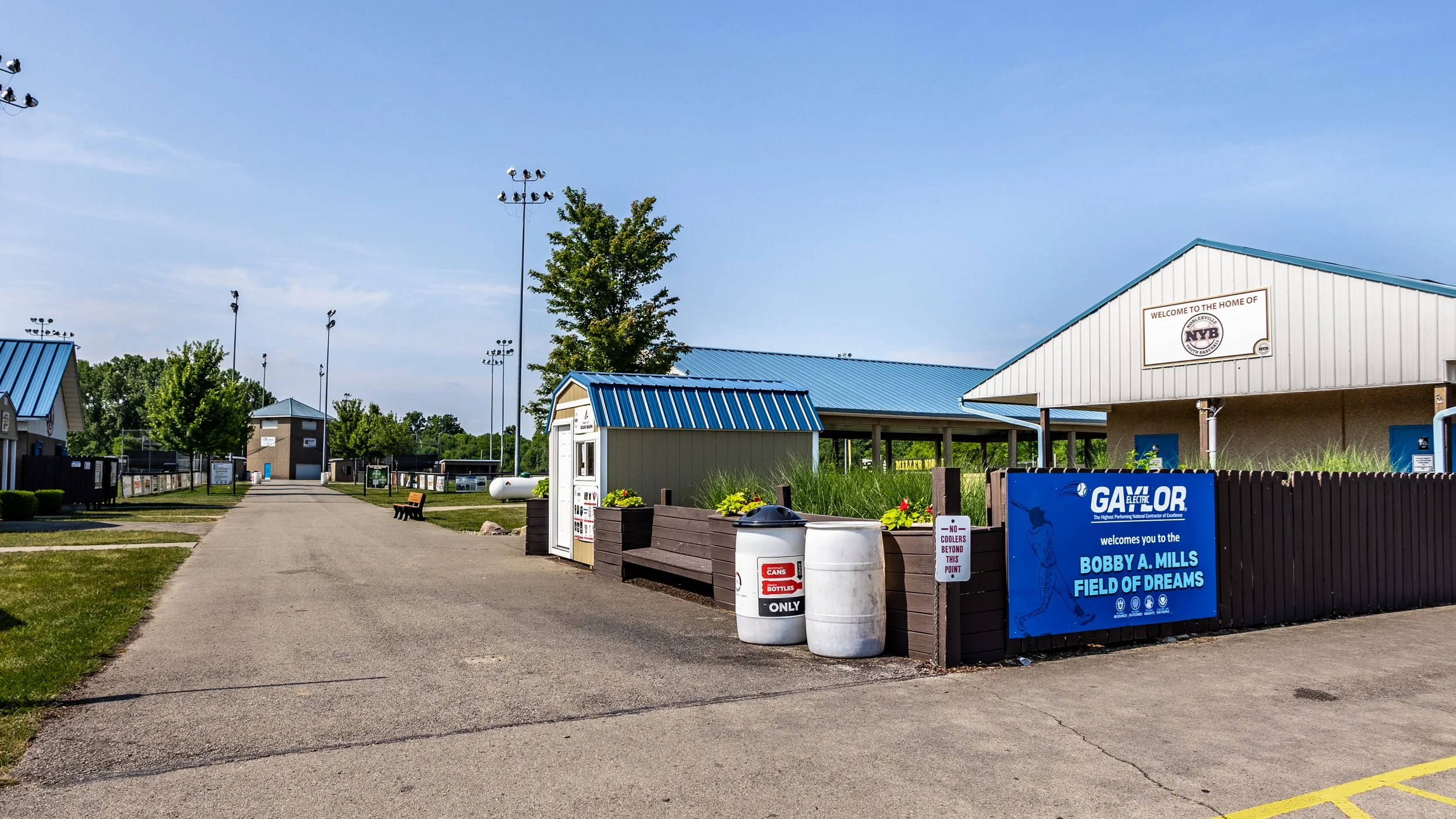 Entrance to Bobby A. Mills Field of Dreams baseball or athletic complex with a sign, pathway, landscaped blue-roofed structures, and tall light poles.