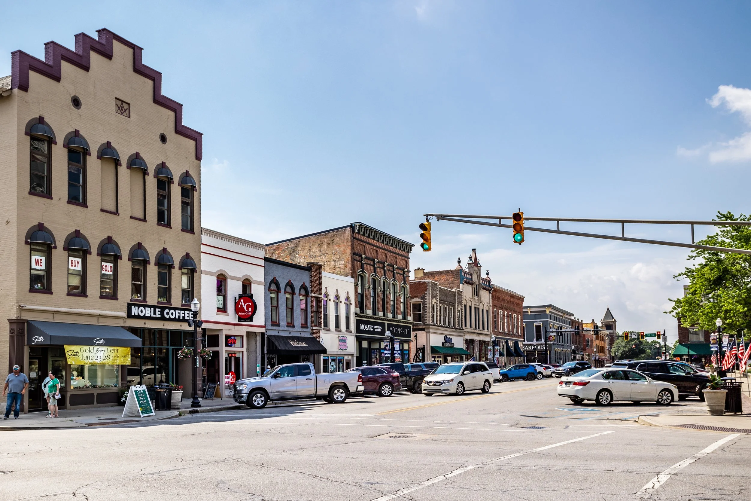 Downtown street with historic buildings, parked cars, and traffic lights on a sunny day.
