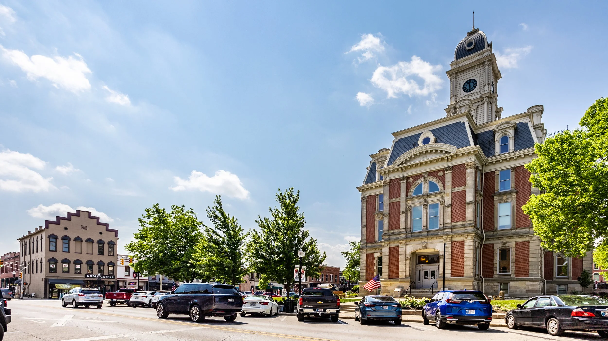 Downtown street with historic courthouse featuring a clock tower, surrounded by trees and parked cars, and a coffee shop building labeled 'Noble Coffee' in the background.