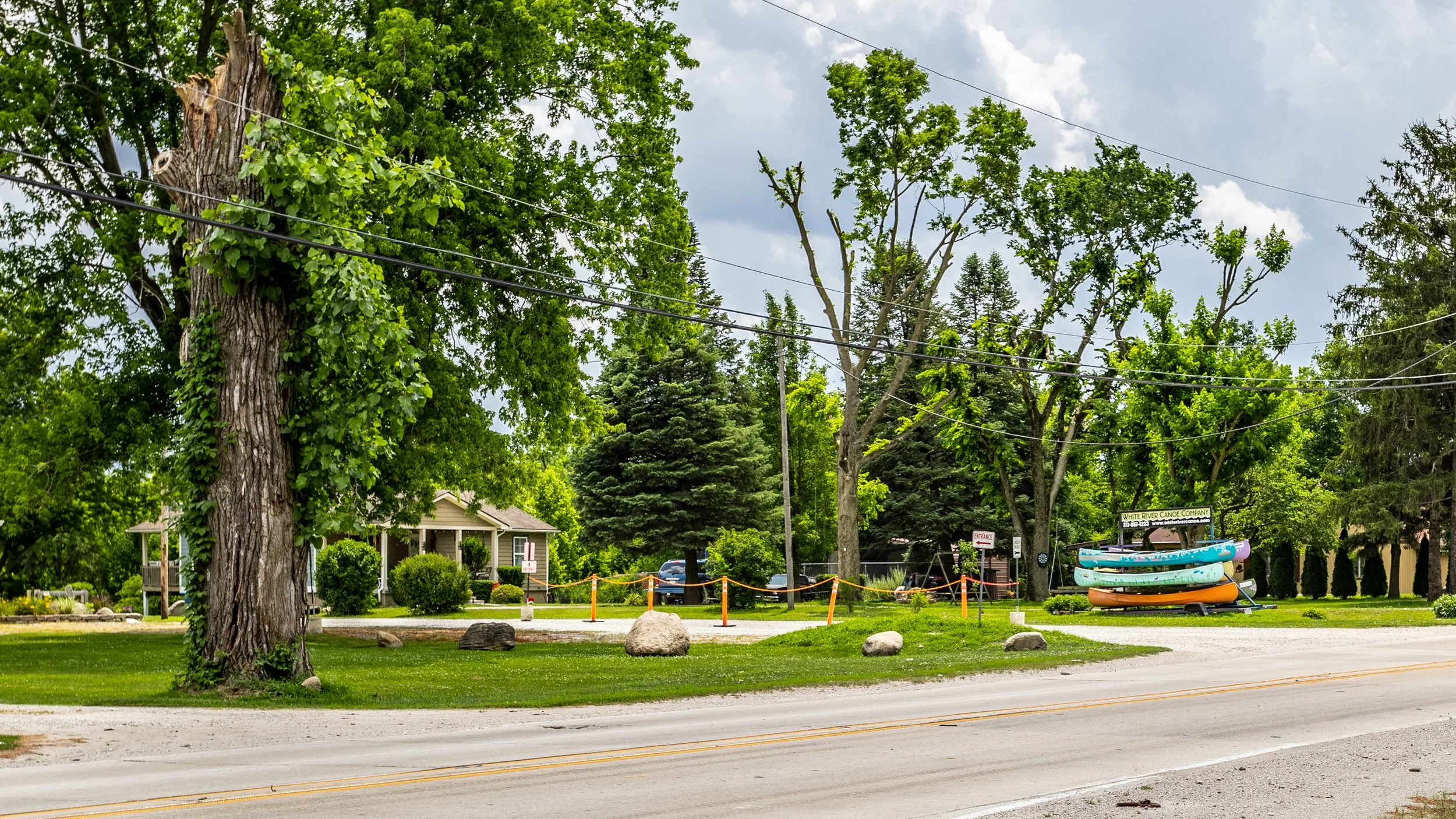 A roadside scene with a large tree, a small house, a row of colorful kayaks on a trailer, and a parking lot, surrounded by green trees and a cloudy sky.