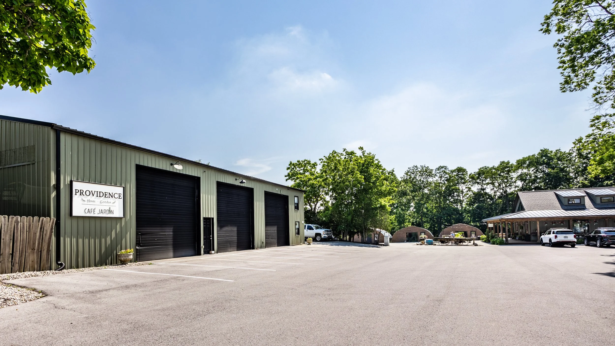 Parking lot outside of a green building with a sign that reads 'Providence House, Café Jardin' and a large patio area with tables, chairs, and a building with a metal roof to the right