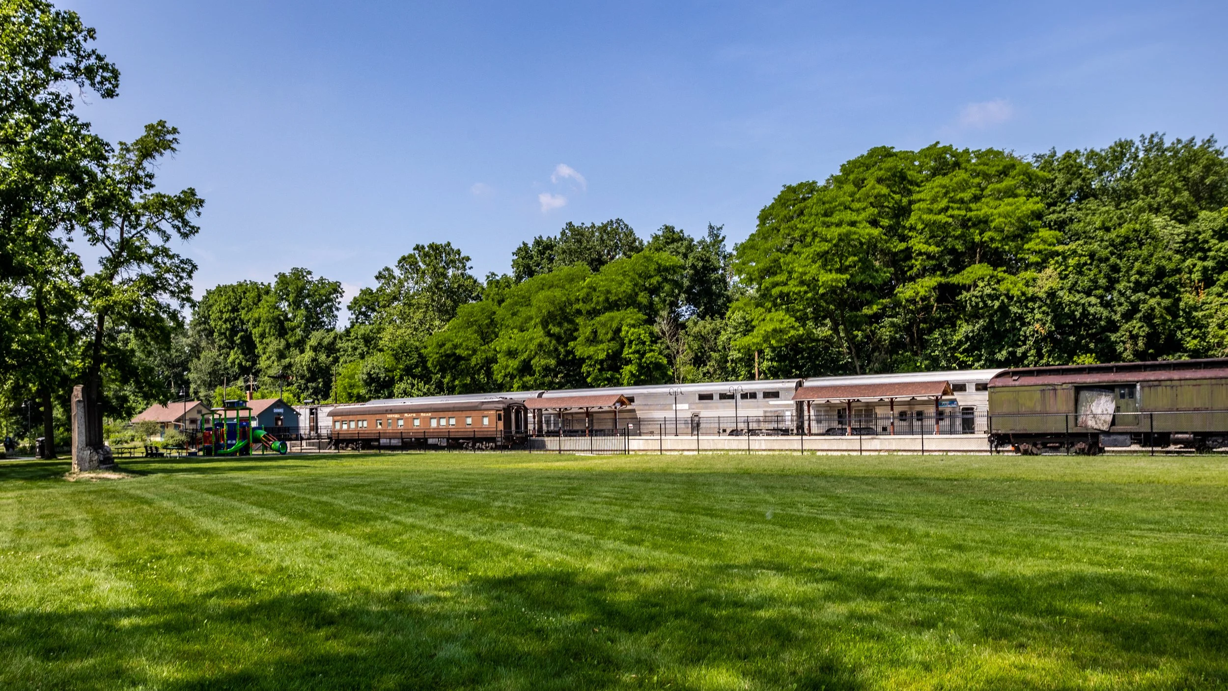 Old train cars and a playground in a park with green grass and trees under a blue sky.
