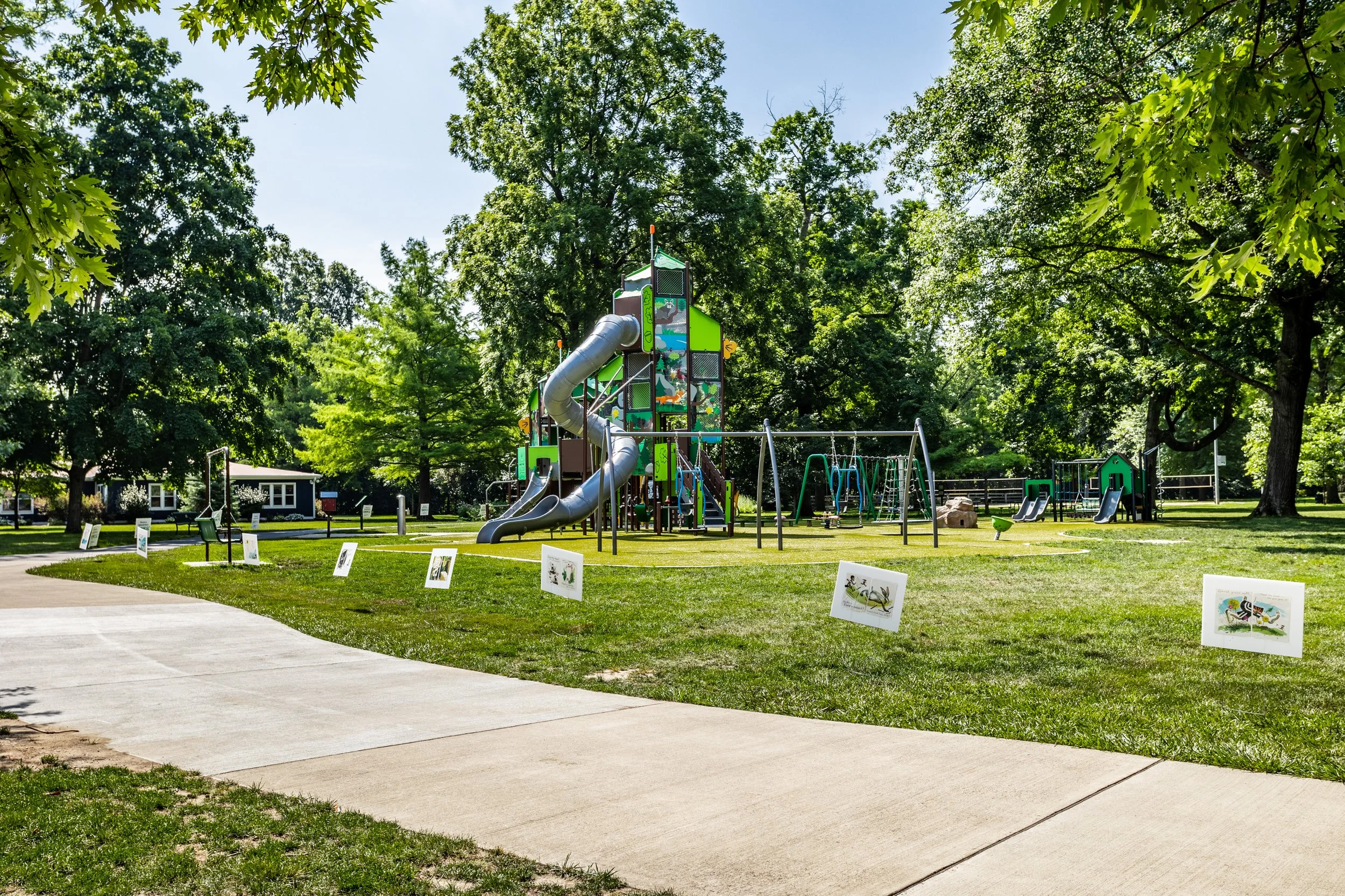 A playground with slides, swing sets, and other equipment surrounded by green grass and tall trees in a park on a sunny day.