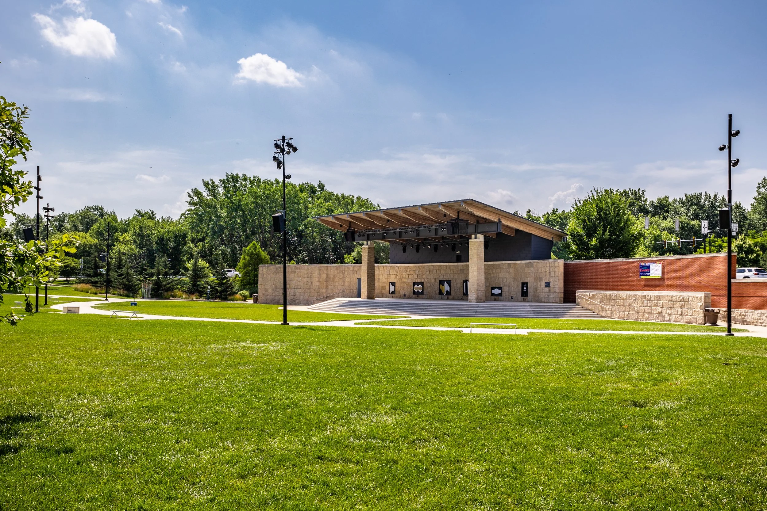 An outdoor stage with a wooden roof and stone pillars, surrounded by a grassy park area with walking paths and trees, under a partly cloudy sky.