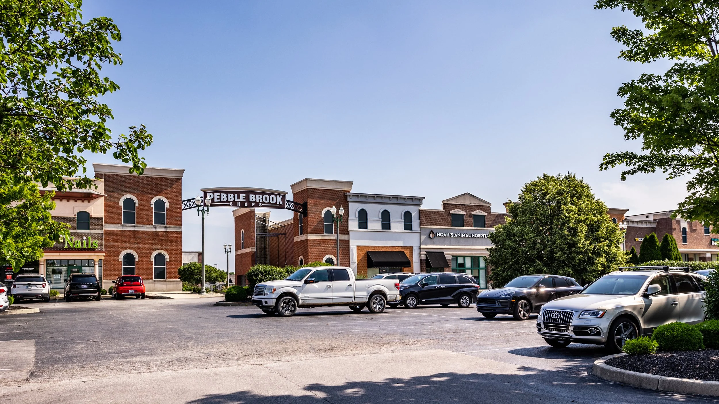 Shopping plaza with storefronts for Nails, Pebble Brook Shops, and Noah's Animal Hospital; cars parked along the street; green trees and a clear blue sky.