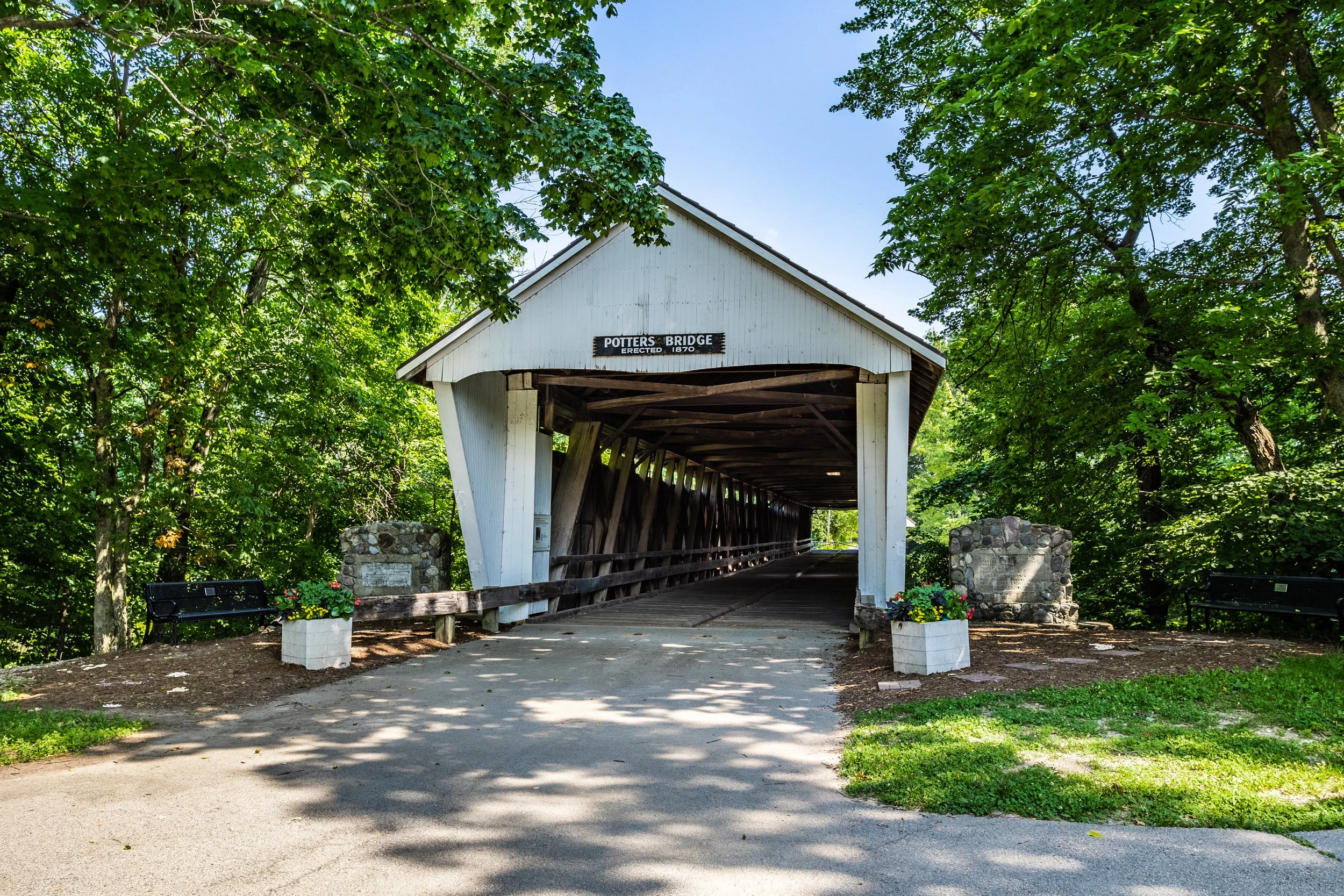 A historic wooden covered bridge called Potters Bridge, built in 1870, surrounded by lush green trees and flowers in planters at the entrance.