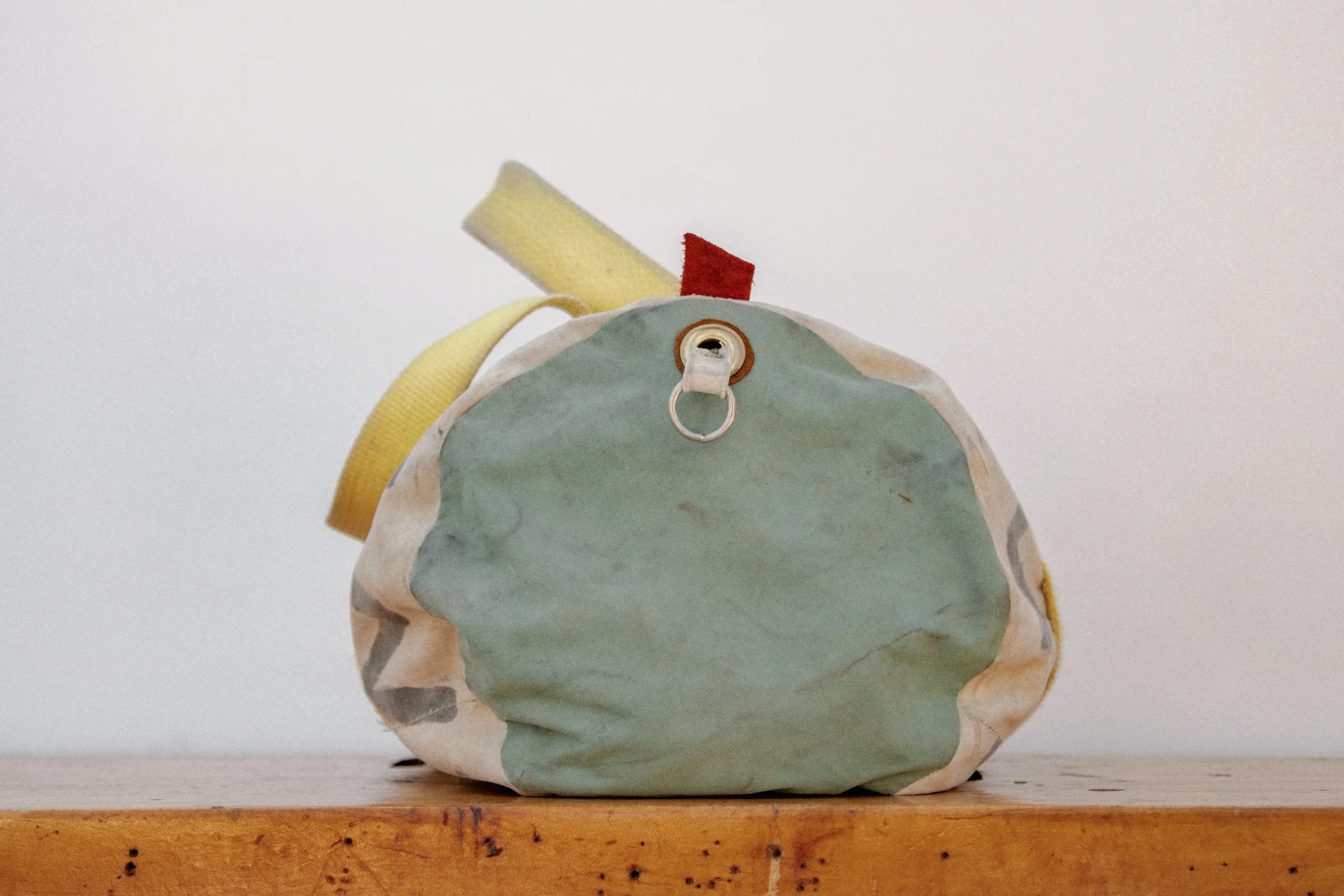 A worn, multicolored fabric bag with a yellow strap, a red tab, and a metal ring attached to the front, resting on a wooden surface against a plain white background.