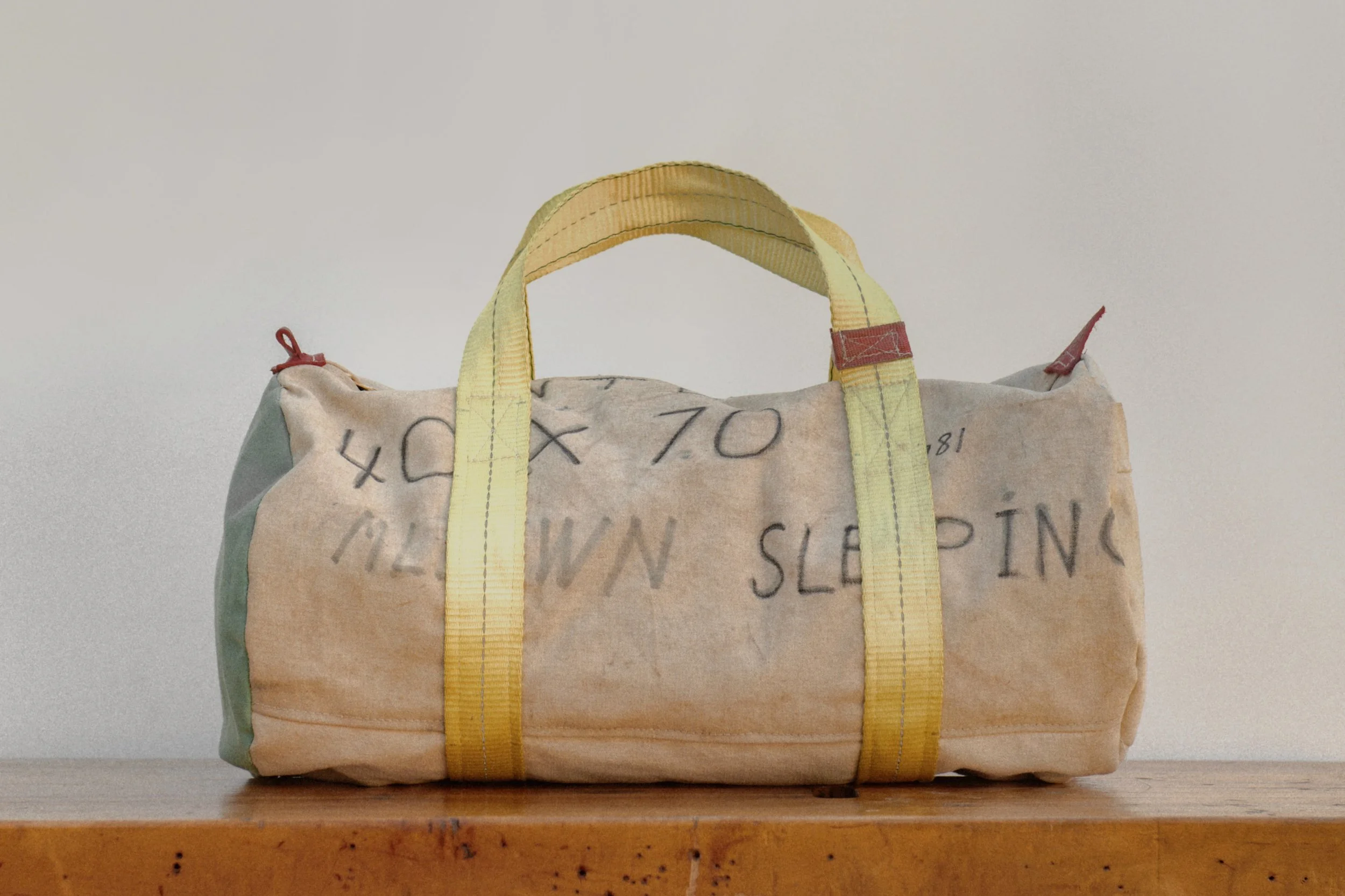 A duffel bag made from a pillowcase with handwritten text, with yellow straps and handles, placed on a wooden surface against a plain background.