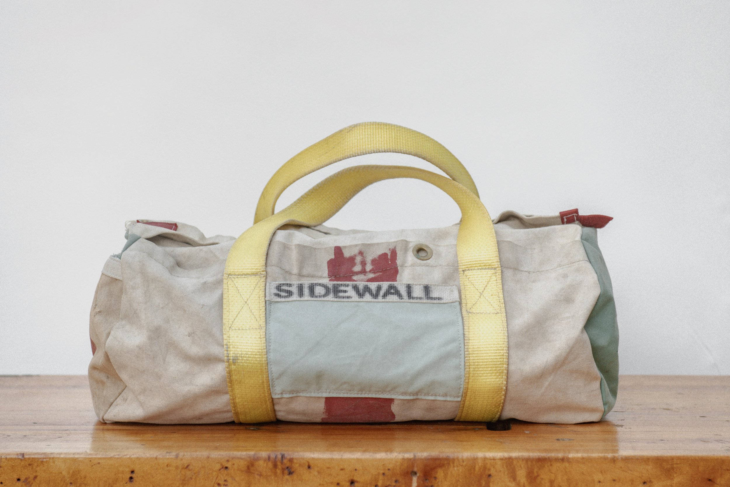 A worn, beige canvas bag with yellow straps and a blue front pocket labeled 'SIDEWALL' sitting on a wooden surface against a plain white wall.