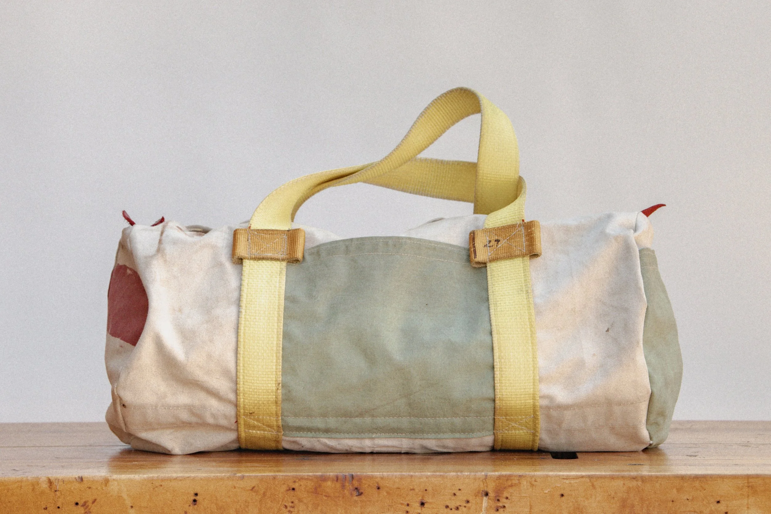 A worn, multicolored duffle bag with yellow straps on a wooden surface against a plain background.