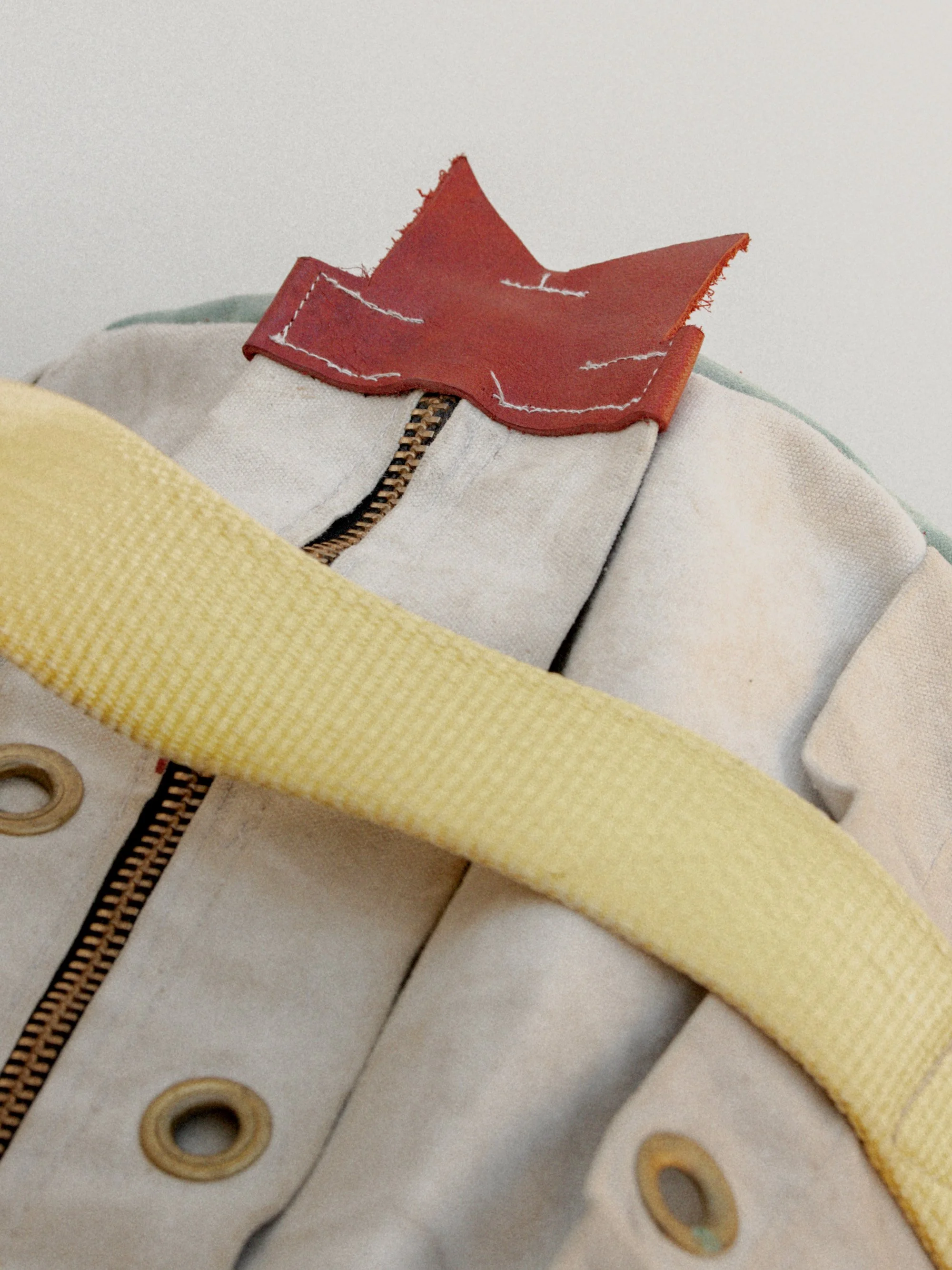 Close-up of a vintage doll's upper body, showing part of its beige outfit with metal eyelets, a yellow strap, and a brown zipper, with a red leather flap at the collar area.
