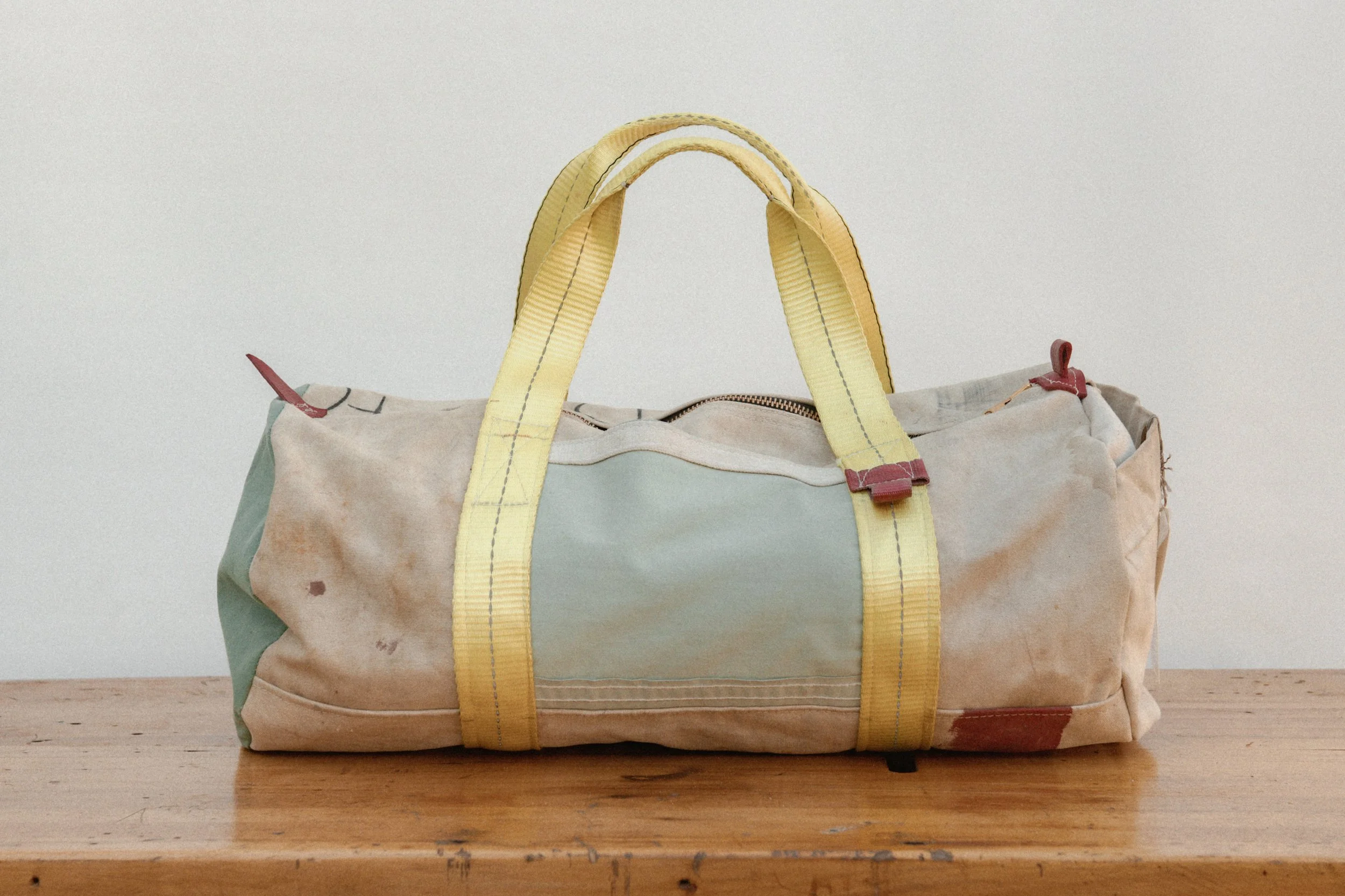 A stained and worn beige and light green duffel bag with yellow handles sitting on a wooden surface against a plain white background.