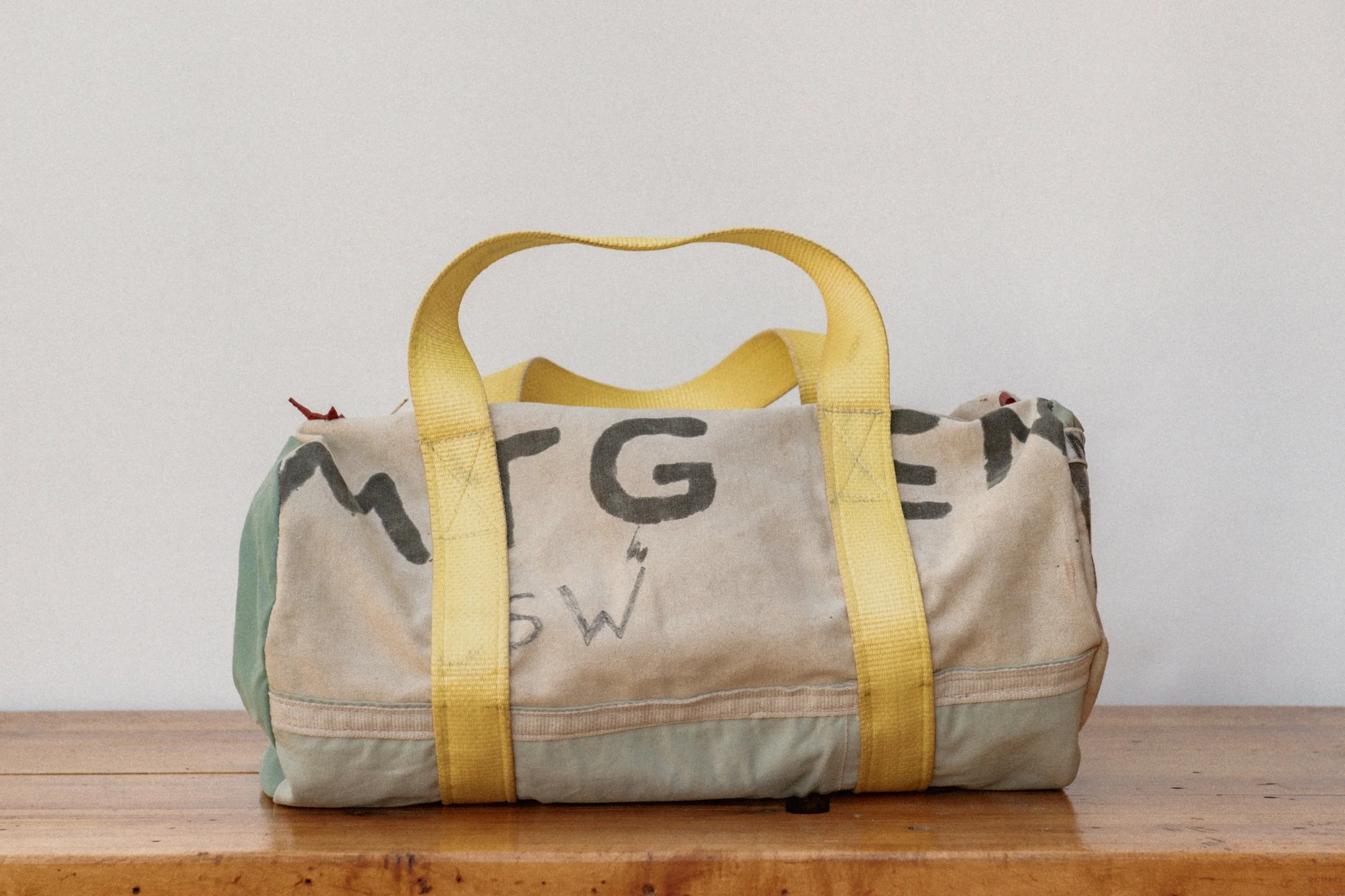 A fabric duffle bag with the words MEETING END with yellow straps and handles placed on a wooden surface against a white background.