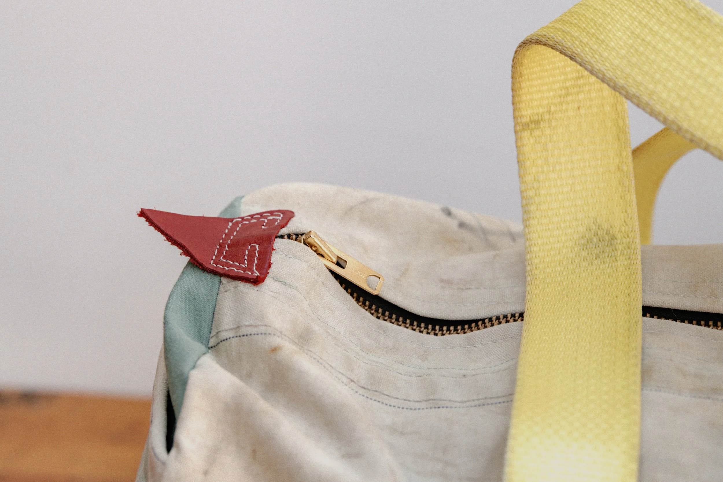 Close-up of a beige fabric bag with a red leather tab and gold zipper, yellow woven strap, and a hint of blue fabric.
