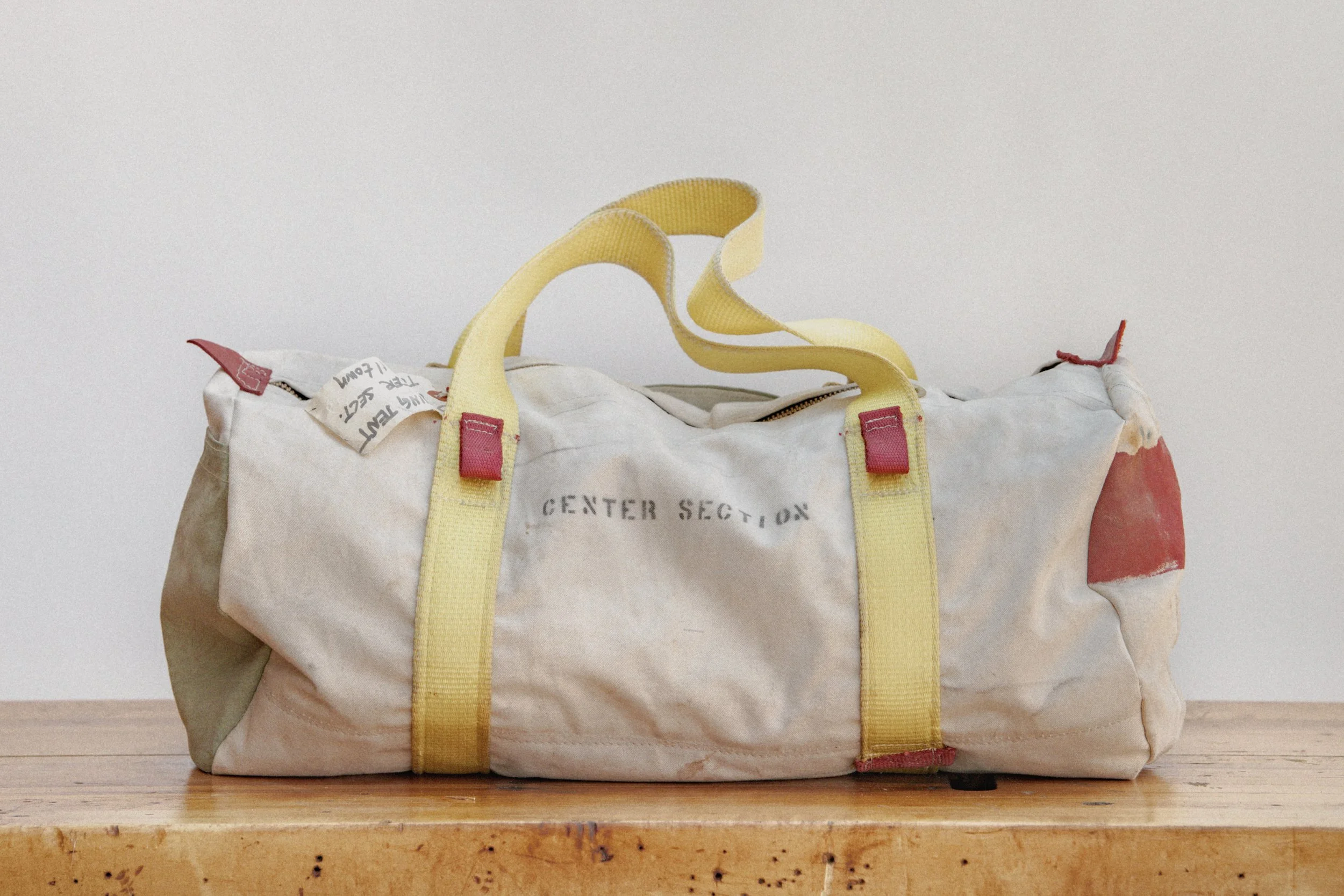 A white duffel bag with yellow straps and red accents, marked with the words 'CENTER SECTION', sitting on a wooden surface against a plain white wall.