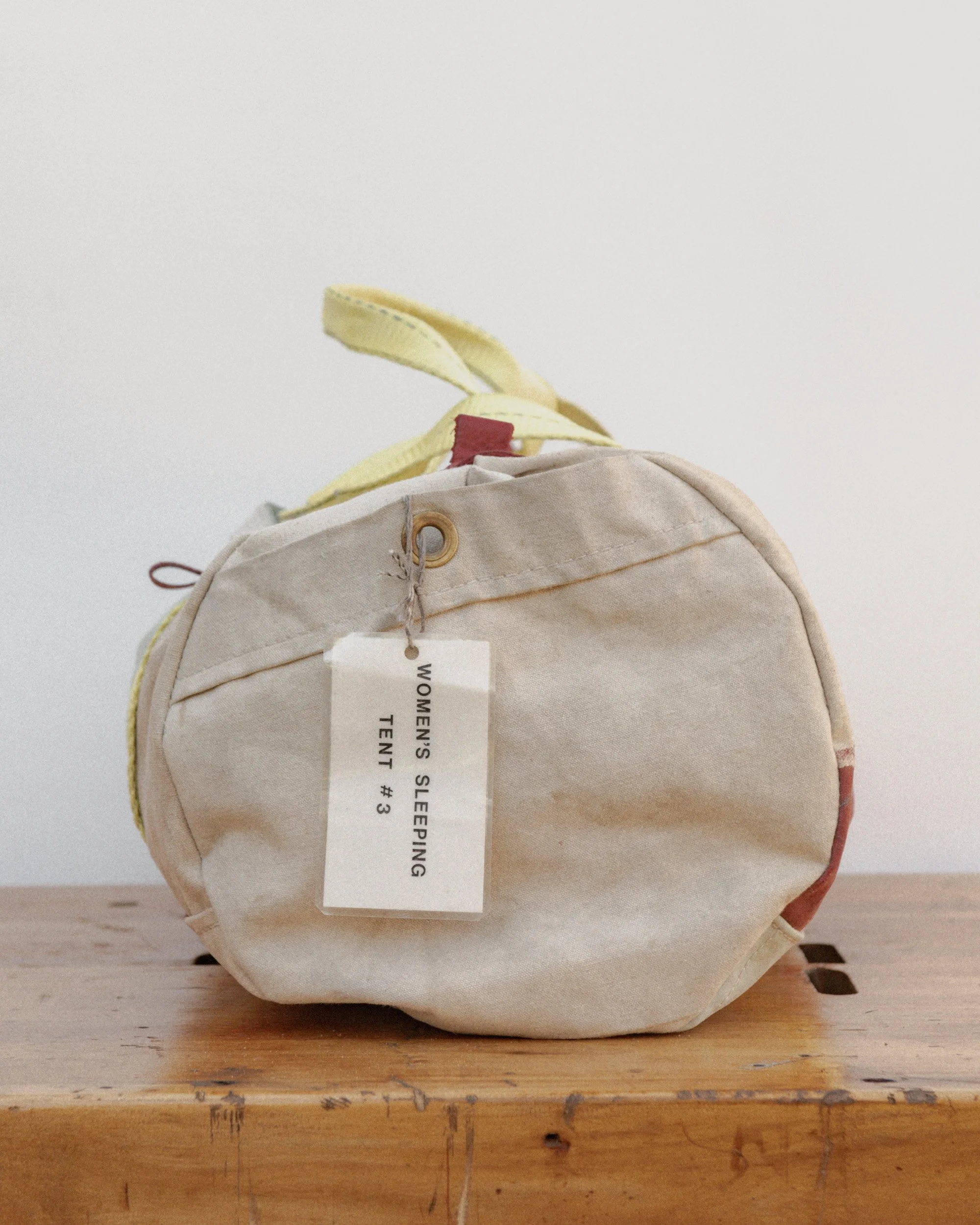 A vintage canvas tote bag with colored handles and a tag that reads 'Women's Sleeping Tent #3', placed on a wooden surface, against a plain white wall.