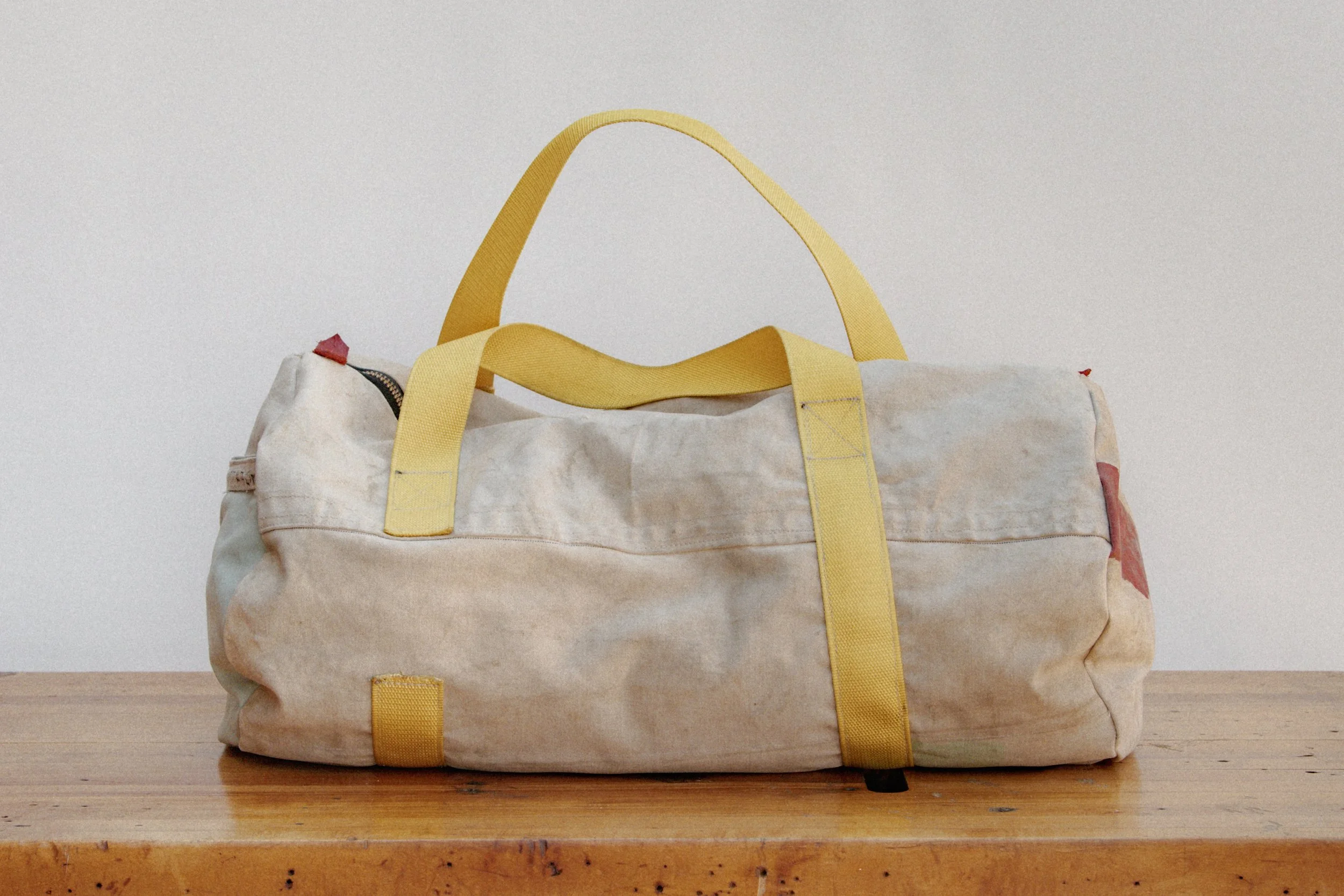 A worn beige duffel bag with yellow straps on a wooden surface against a plain white background.