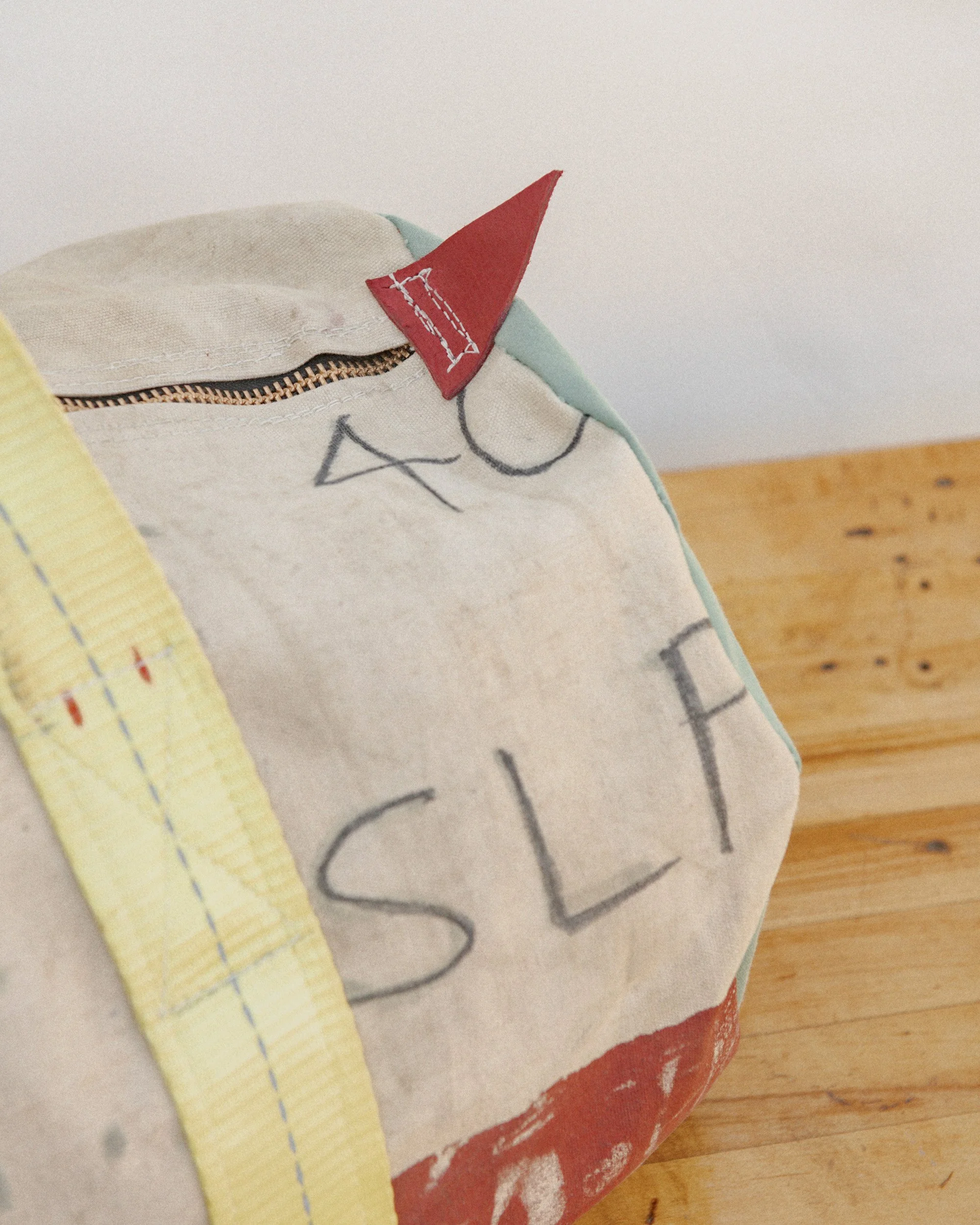 Close-up of a fabric bag with a red triangle sewn near the zipper, hand-drawn letters spelling "SLA" on the fabric, a yellow ruler partially visible on the left, and a wooden surface in the background.