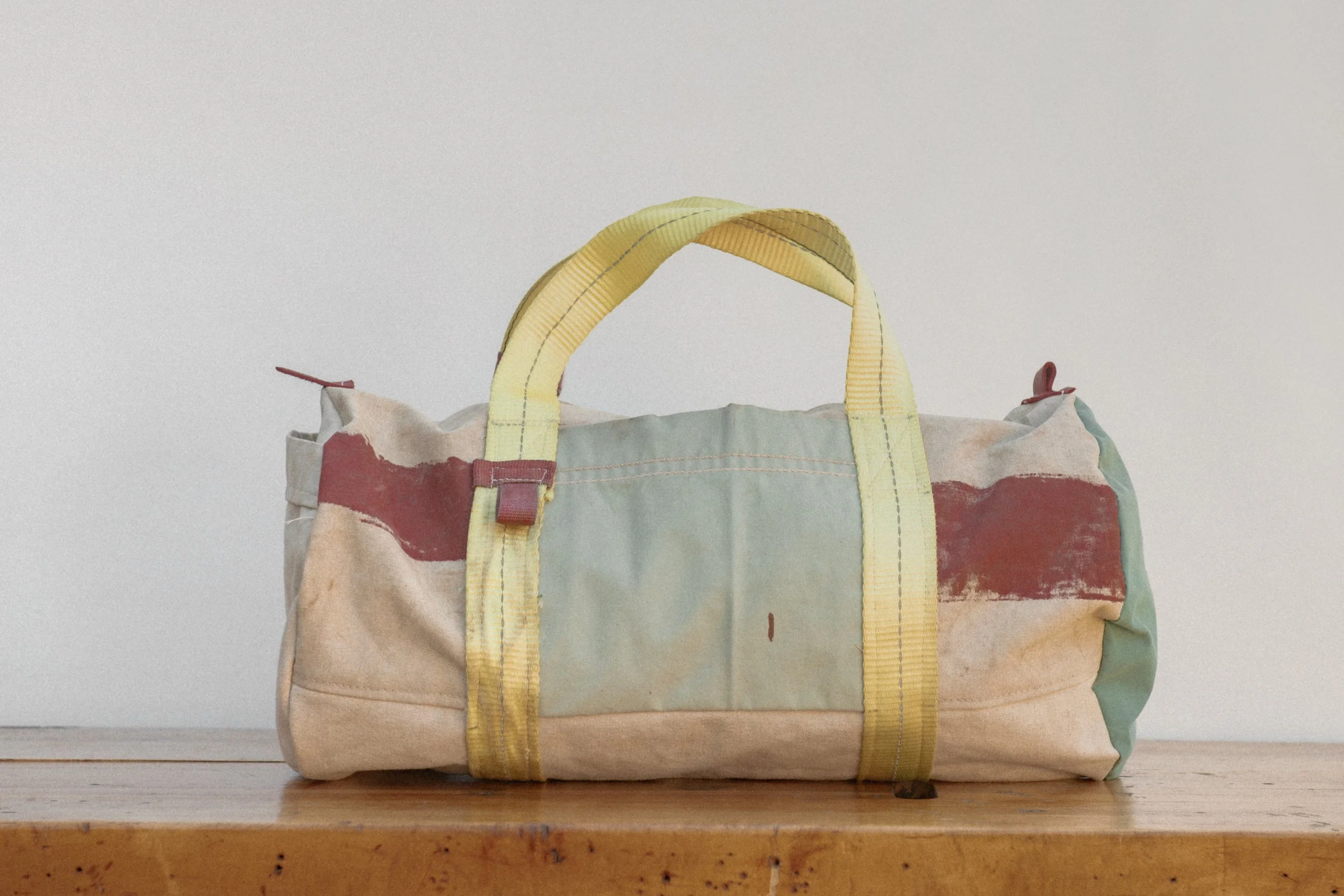 A worn duffel bag with yellow handles and multicolored fabric, sitting on a wooden surface in front of a plain wall.