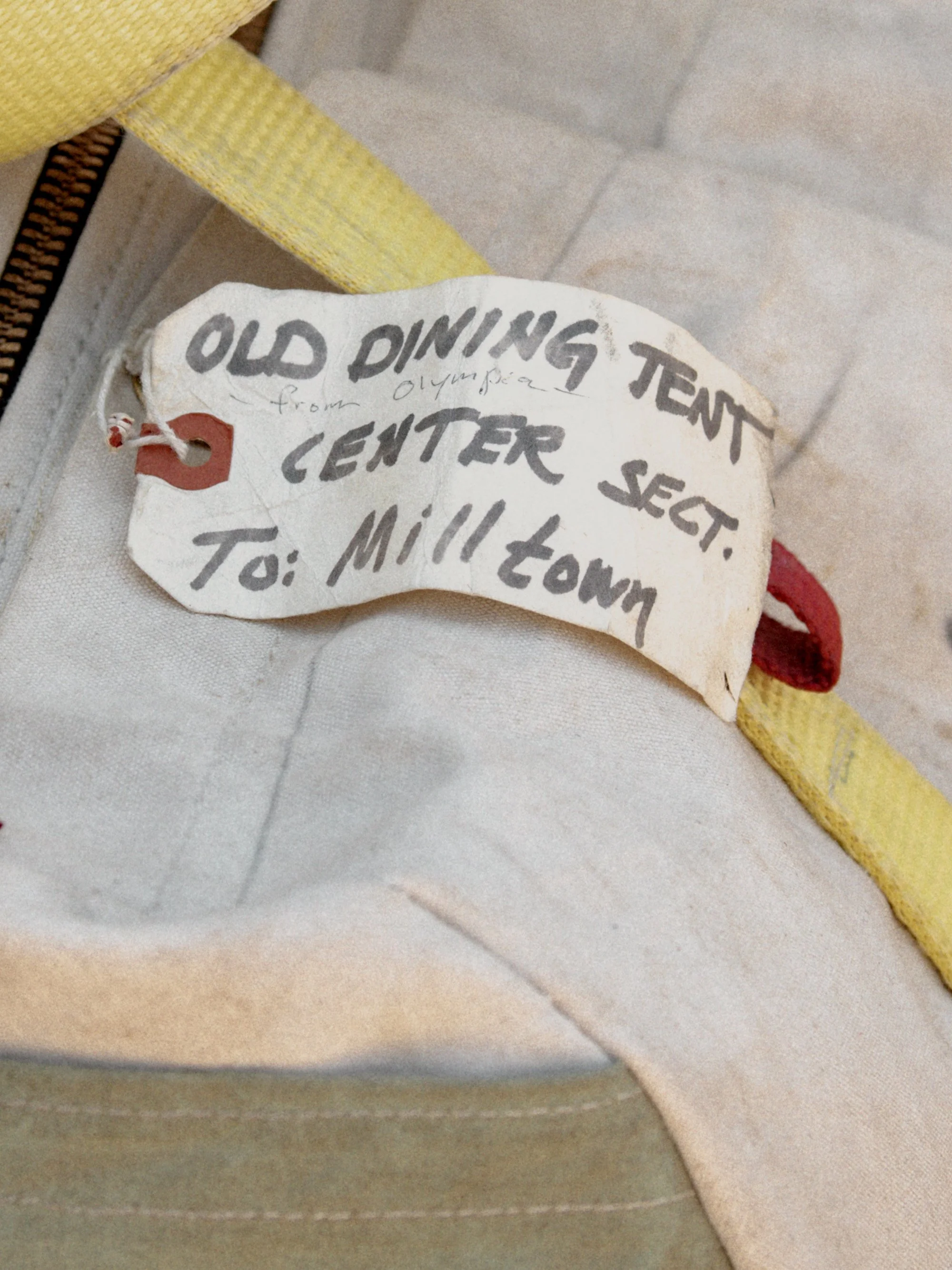 A cloth tag with handwritten text attached to a yellow and beige fabric item, indicating it is an old diving tent from the Olympia Center section, addressed to Milltown.