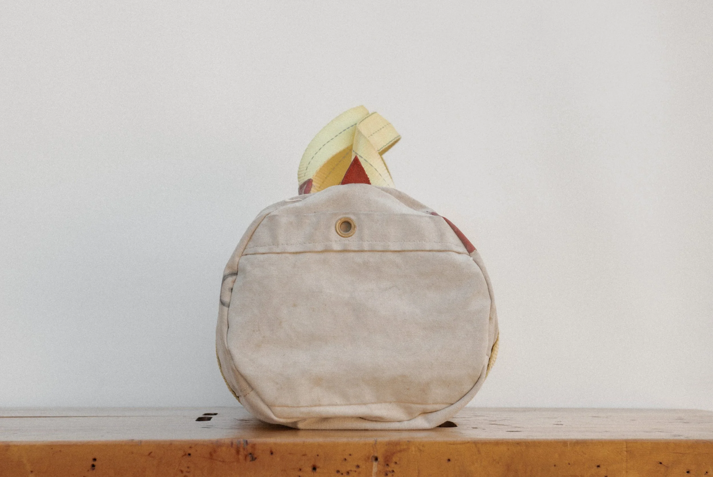A worn beige backpack resting on a wooden surface against a plain white wall, with a yellow and red item partially visible inside.