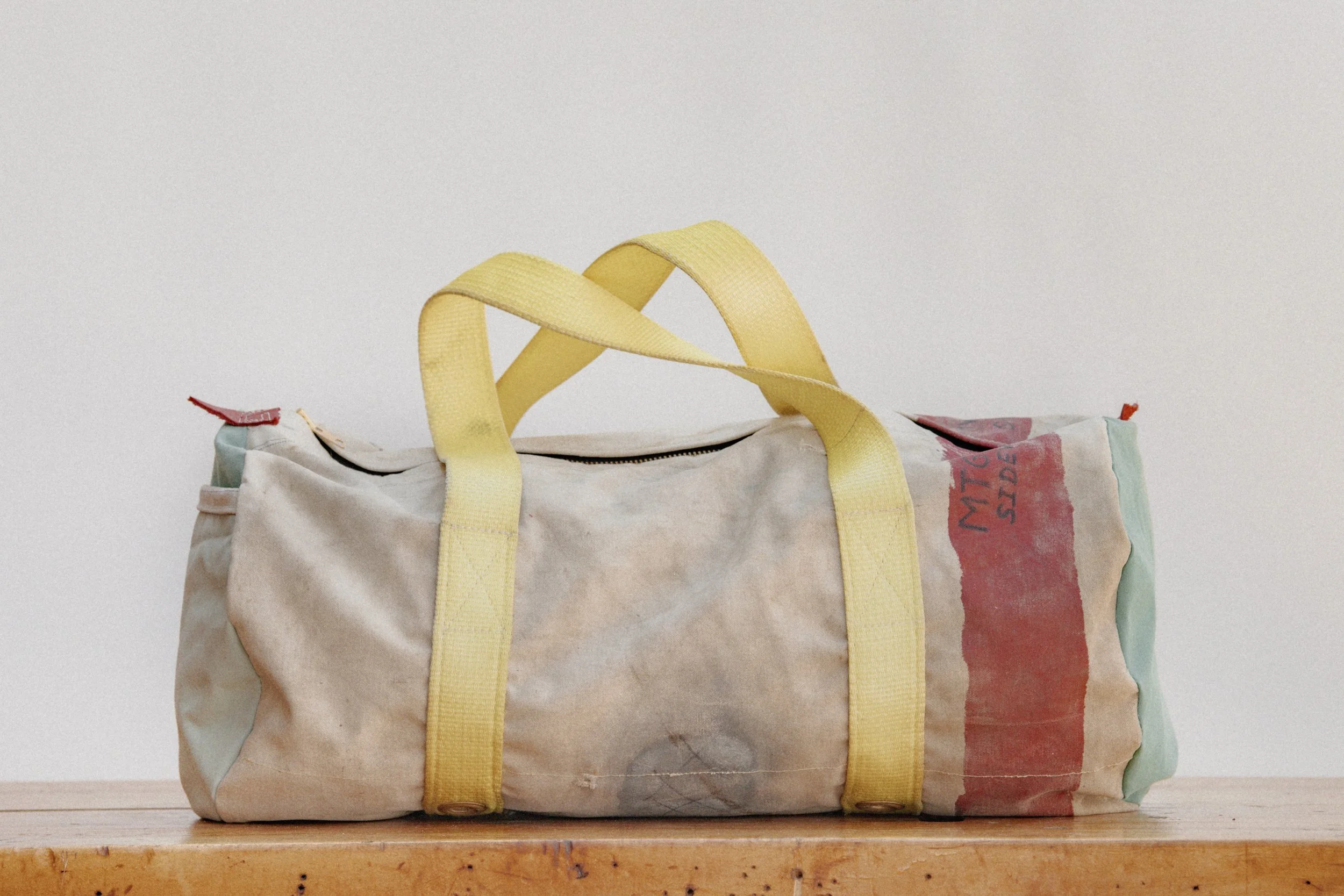 A vintage duffel bag made of worn, faded fabric with yellow straps, resting on a wooden surface against a plain white wall.
