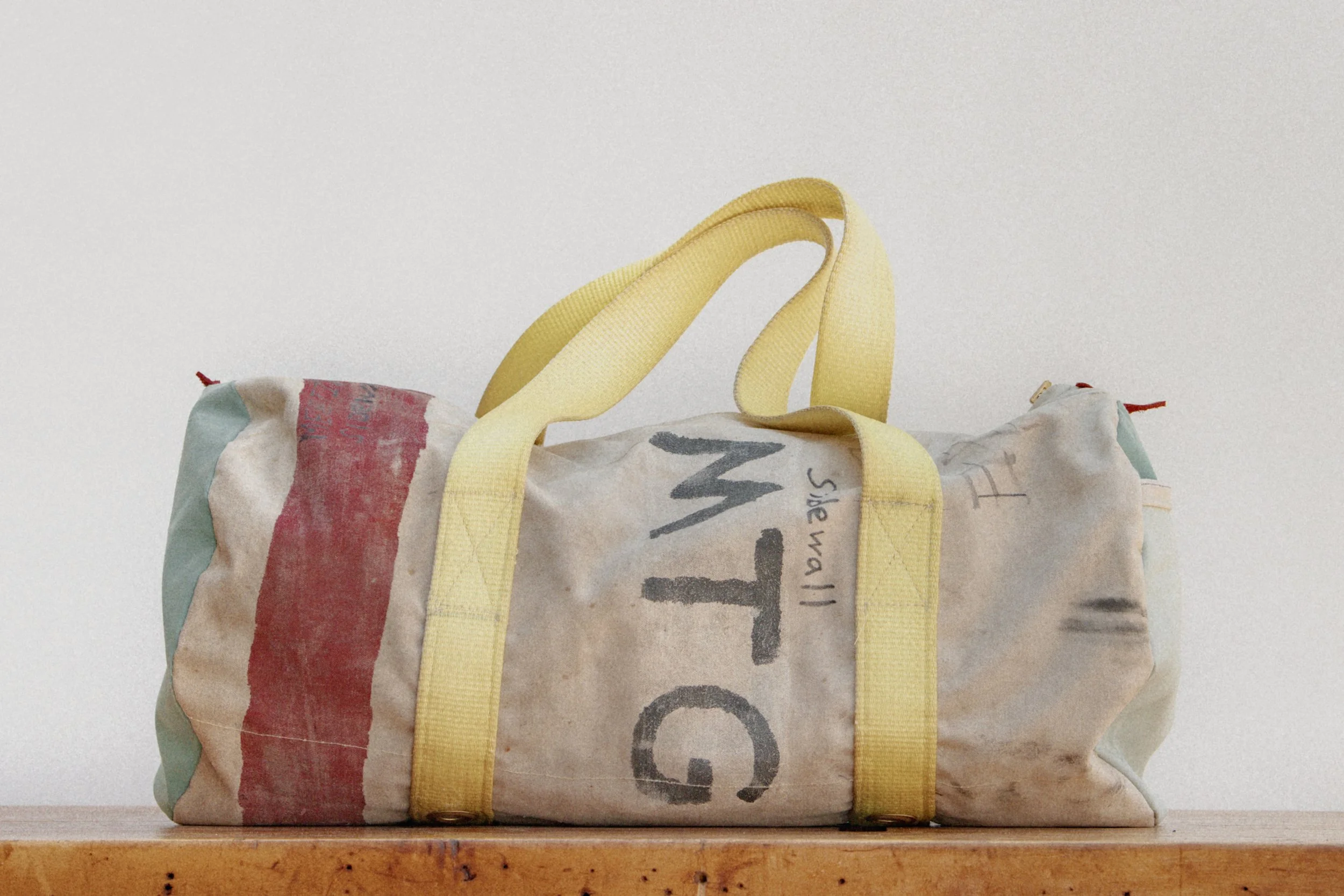 A worn canvas duffel bag with yellow handles and straps, resting on a wooden surface against a plain white background.