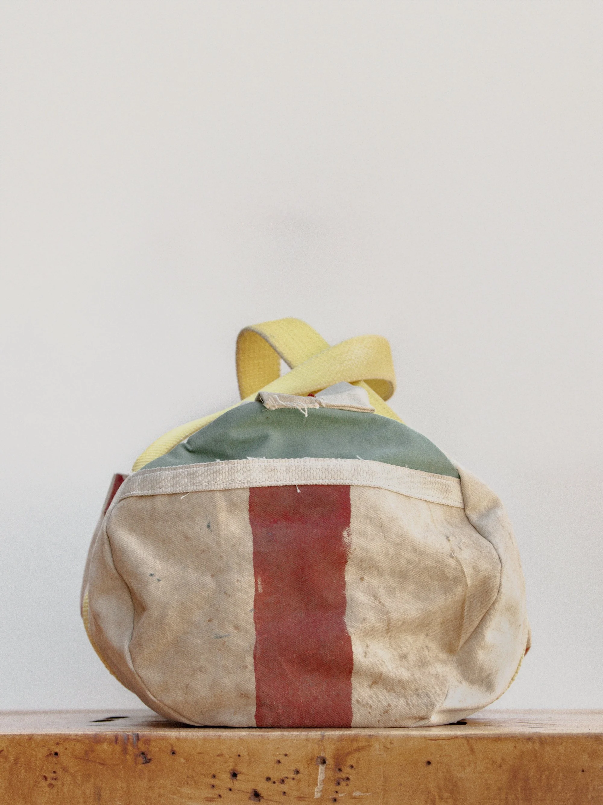 A worn, multicolored canvas bag with a vertical red stripe, sitting on a wooden surface against a plain white background.