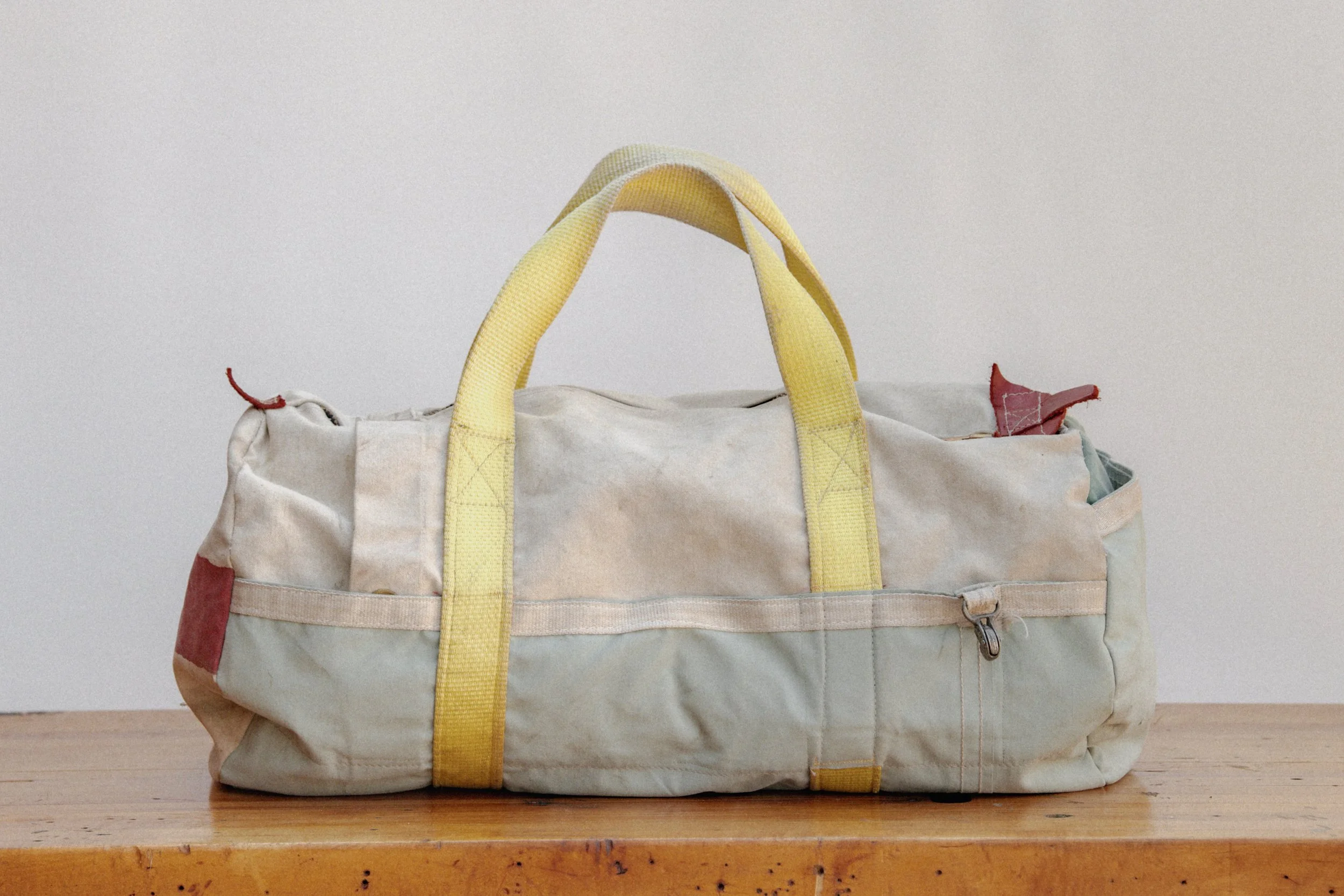 A worn, off-white duffel bag with yellow handles resting on a wooden surface against a plain white wall.