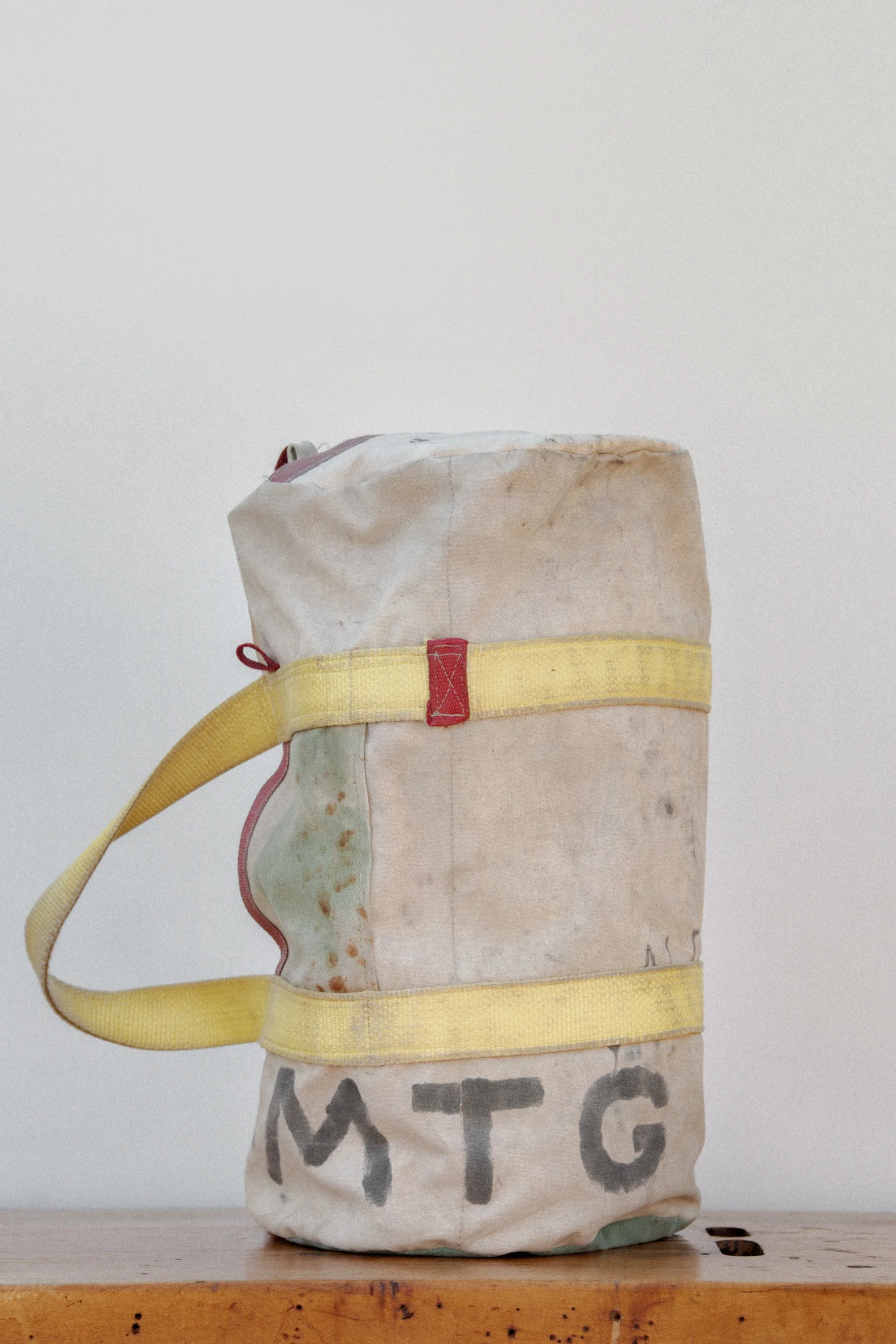 A white cloth bag with yellow straps and red accents, marked with the letters 'MTG' in black, placed on a wooden surface against a plain background.