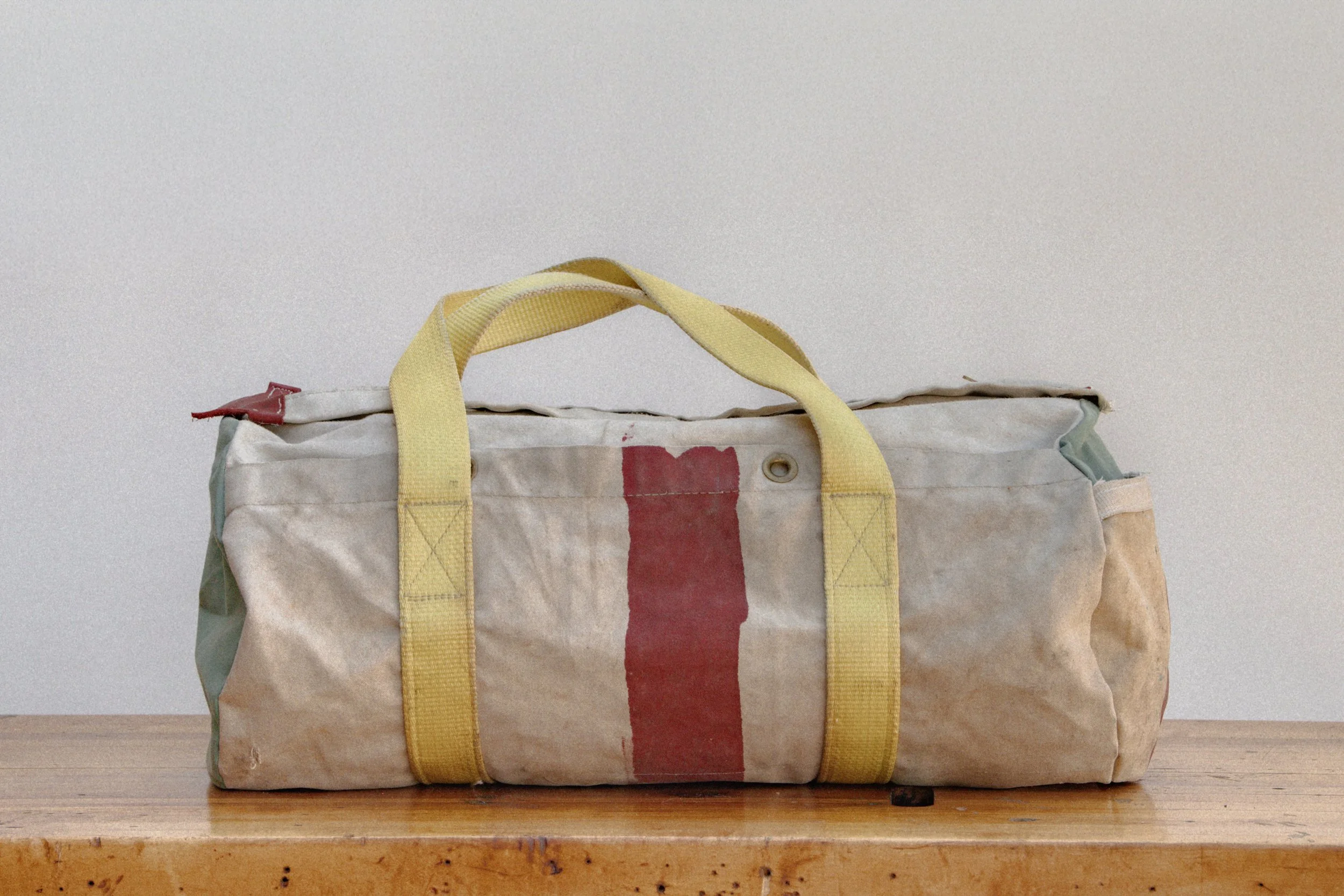 A worn, beige fabric duffel bag with yellow straps and a red stripe in the middle, sitting on a wooden surface against a plain white background.