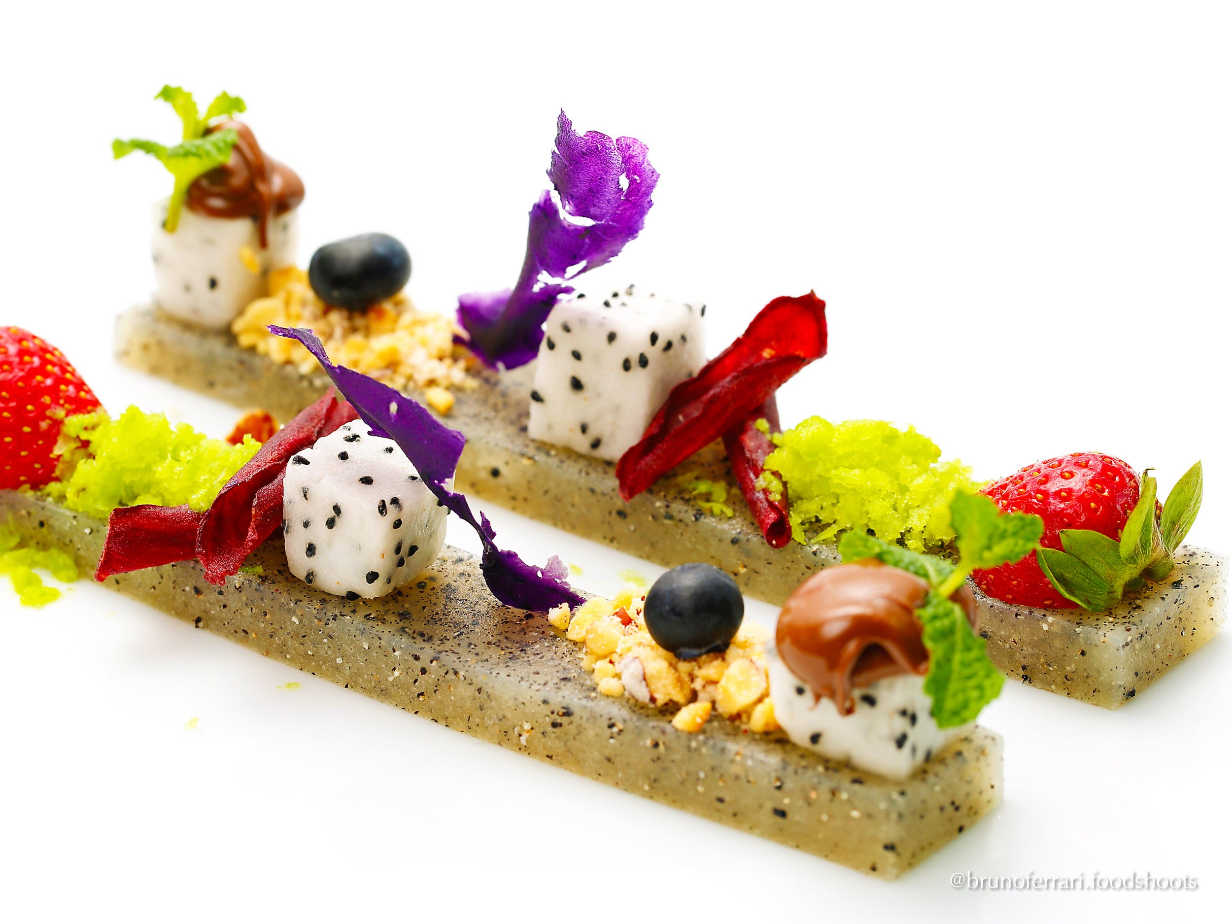 Deconstructed dessert with colorful fruits, edible flowers, and various toppings on two rectangular textured base layers, garnished with seeds, nuts, and mint leaves, on a white background.