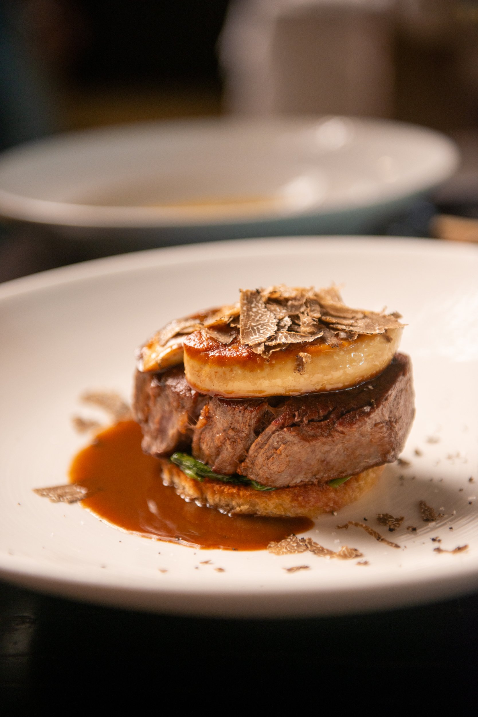 A plated gourmet beef dish topped with foie gras and black truffle shavings, with sauce on the side. beef tenderloin black angus rossini